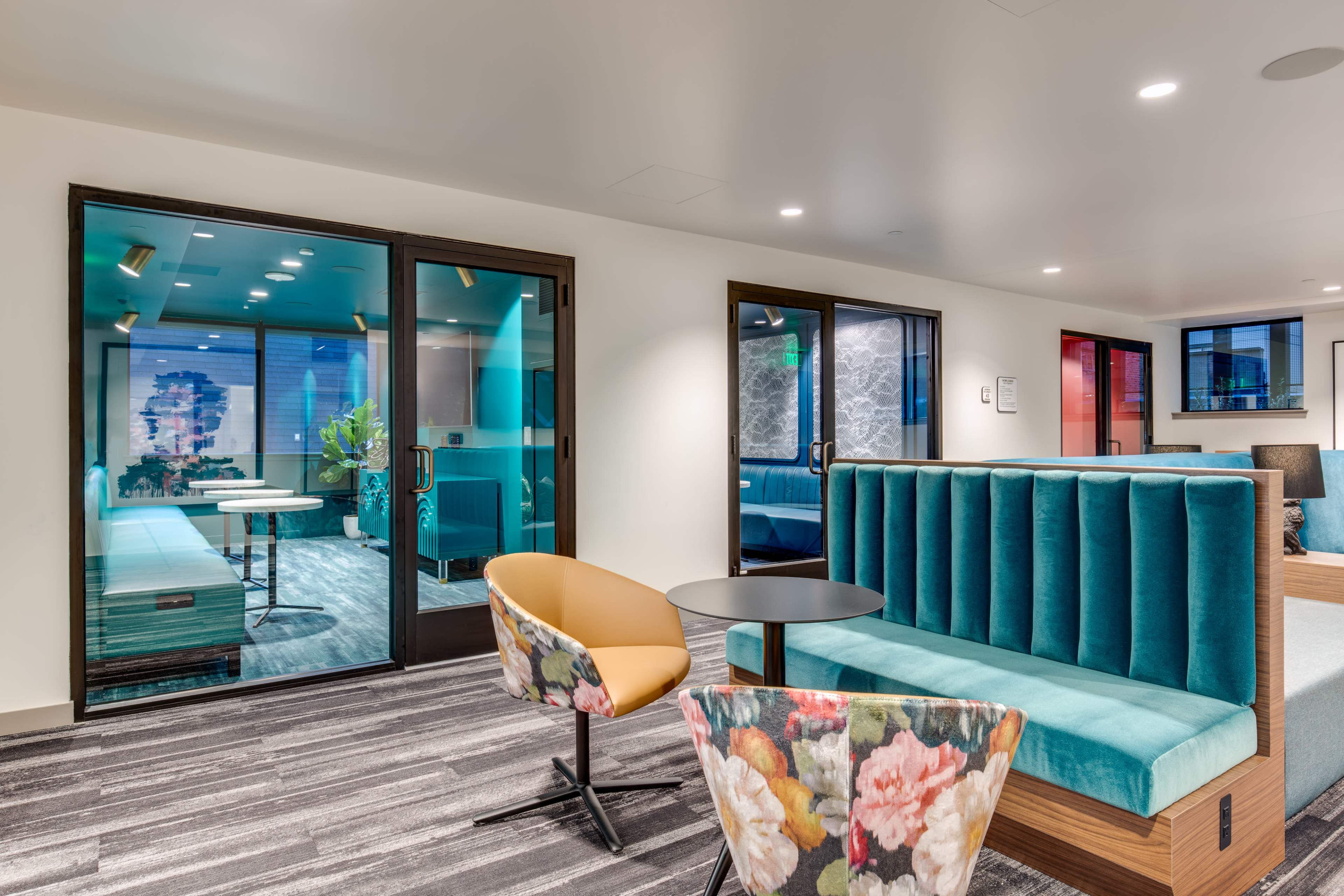 A modern lounge area with teal seating, a small black table, and glass doors leading to another brightly lit room.