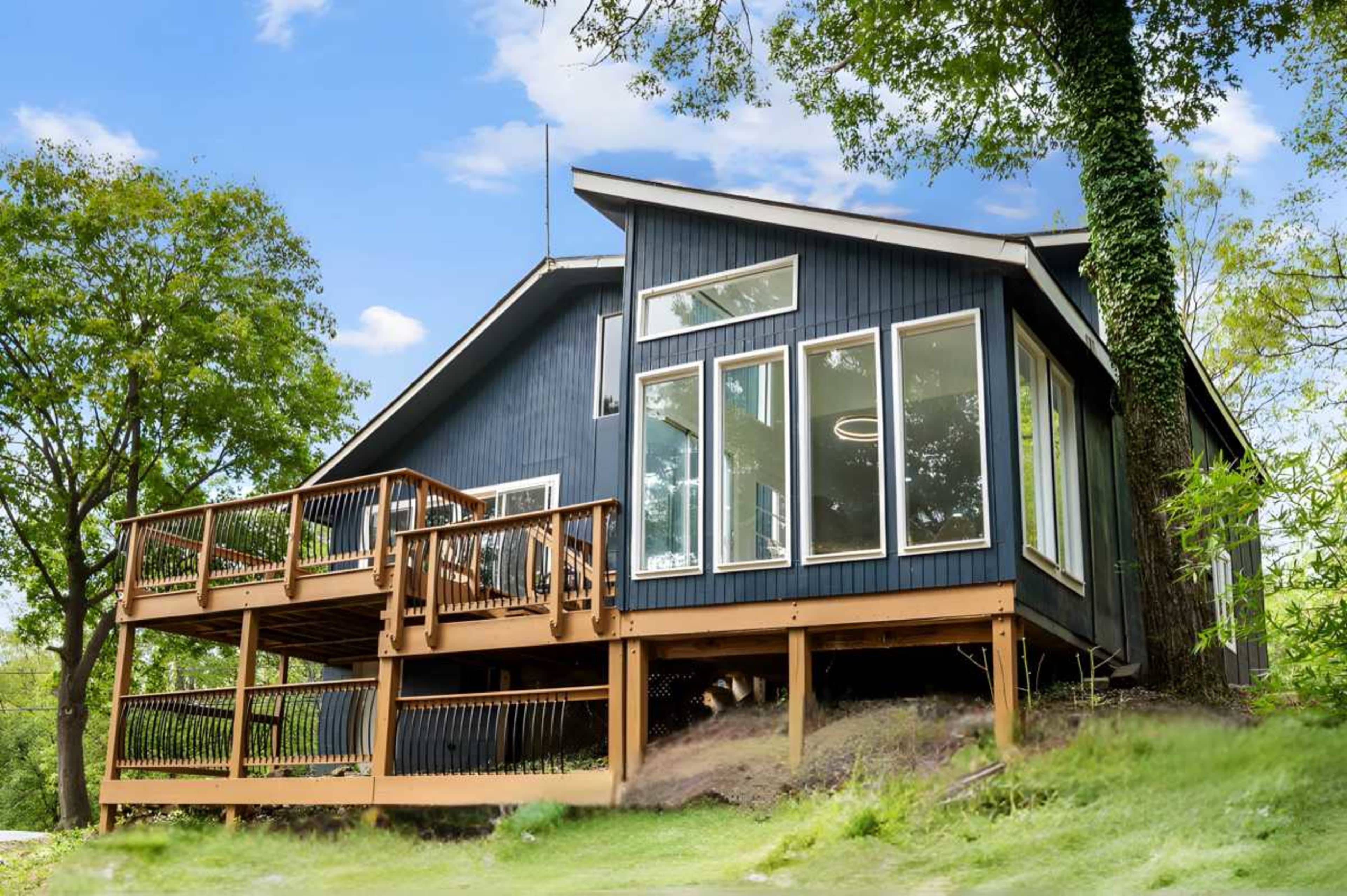 A modern blue house with large windows and a wooden deck is situated on a grassy slope surrounded by trees.