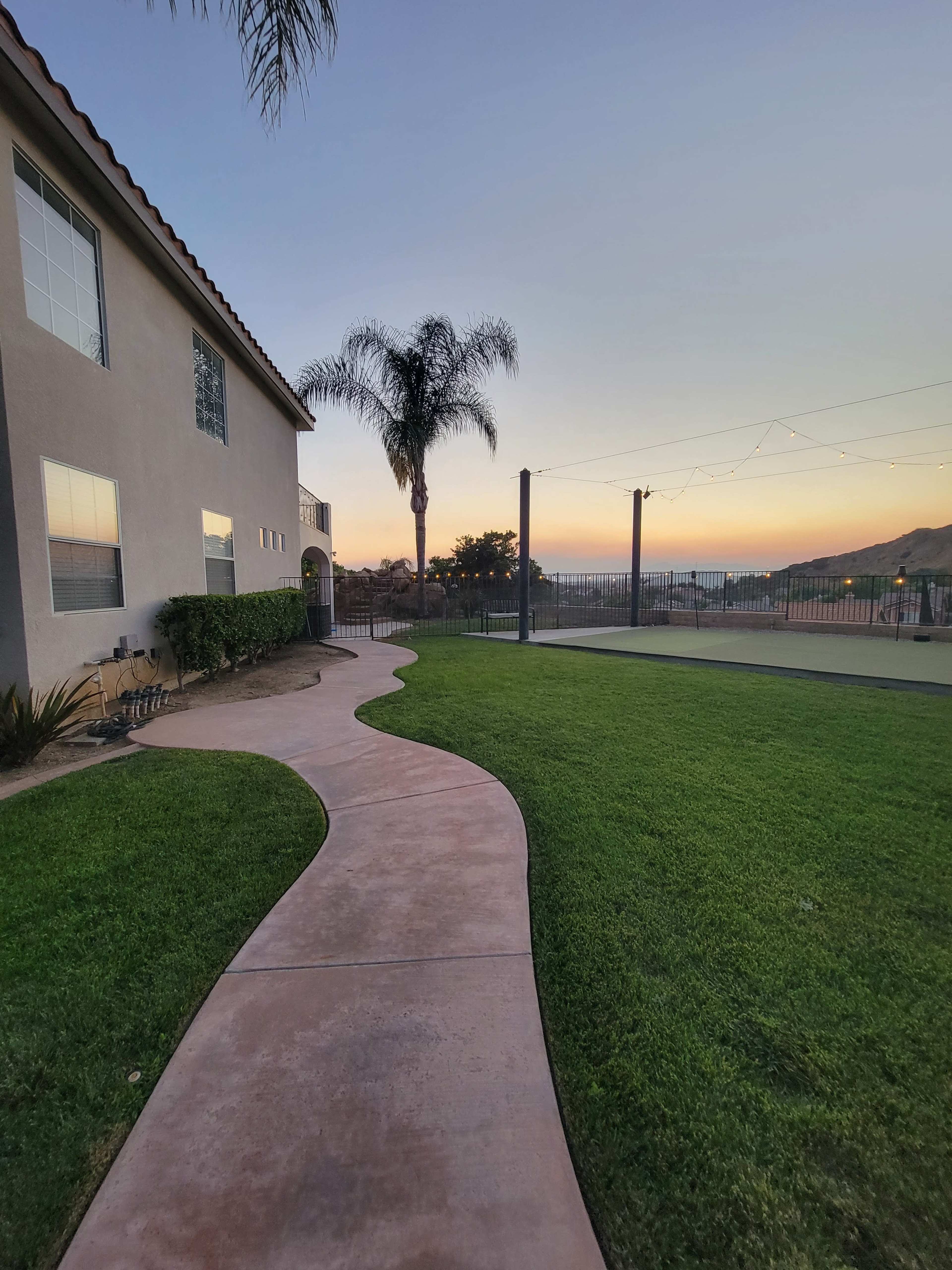 A concrete walkway curves through a green lawn beside a house, leading towards a fenced area with a sunset backdrop.