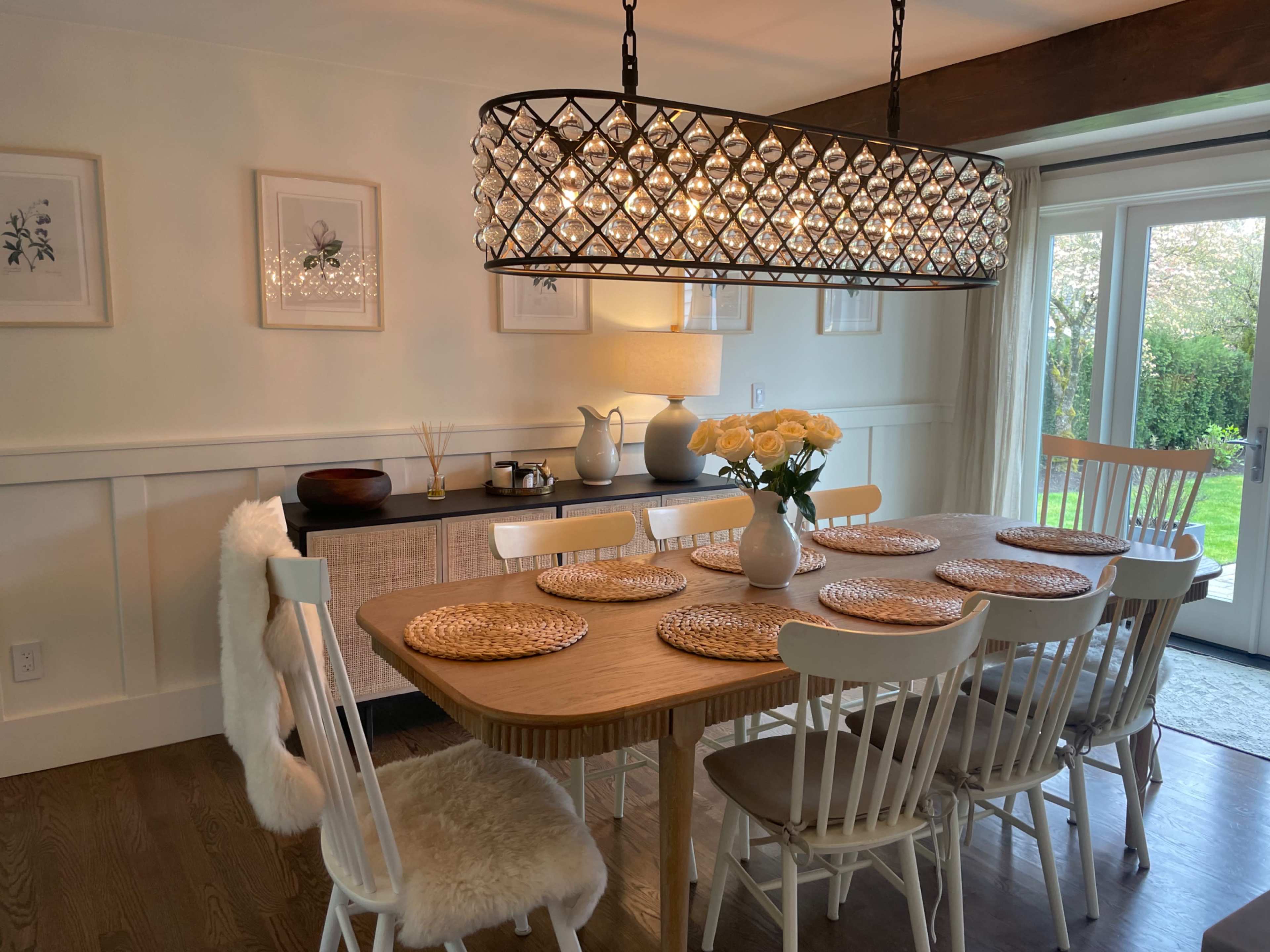 A modern dining room features a large wooden table surrounded by white chairs, with a crystal chandelier overhead and a vase of flowers in the center.