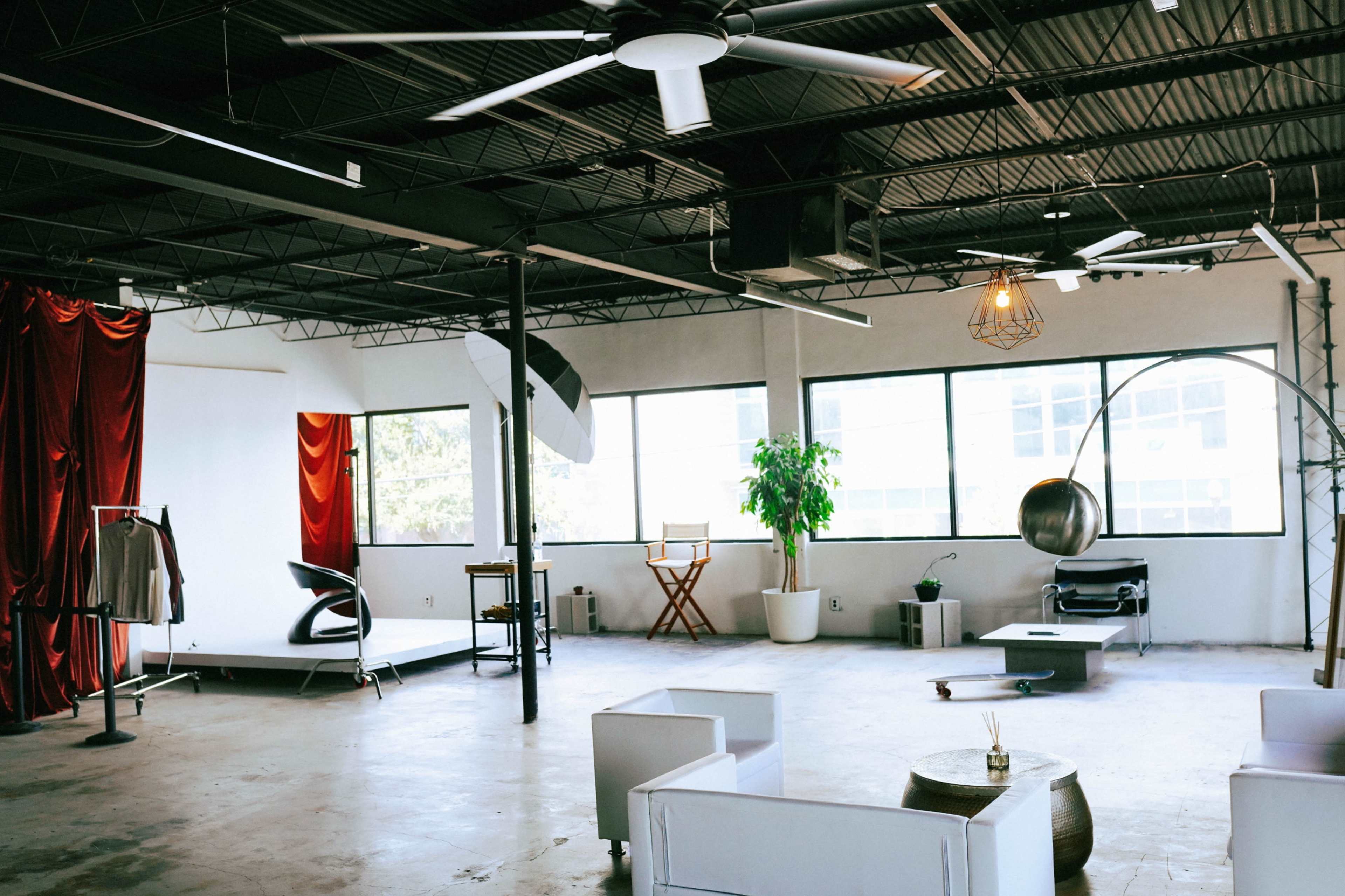 The image shows a spacious, minimalist studio with exposed ceilings, large windows, a stage area, and various seating arrangements.