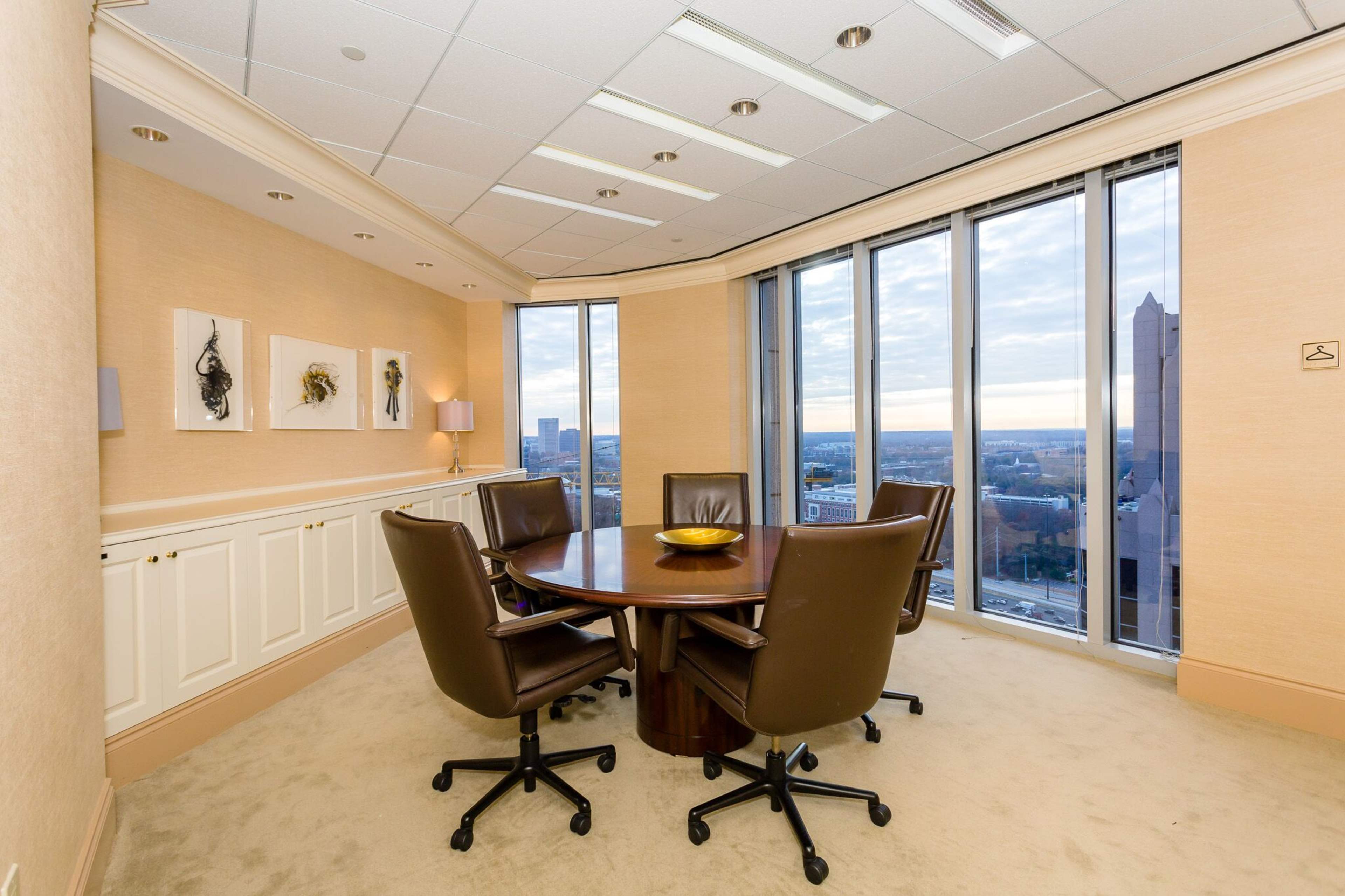 A meeting room features a round table surrounded by four leather chairs, with large windows offering a view of the cityscape.