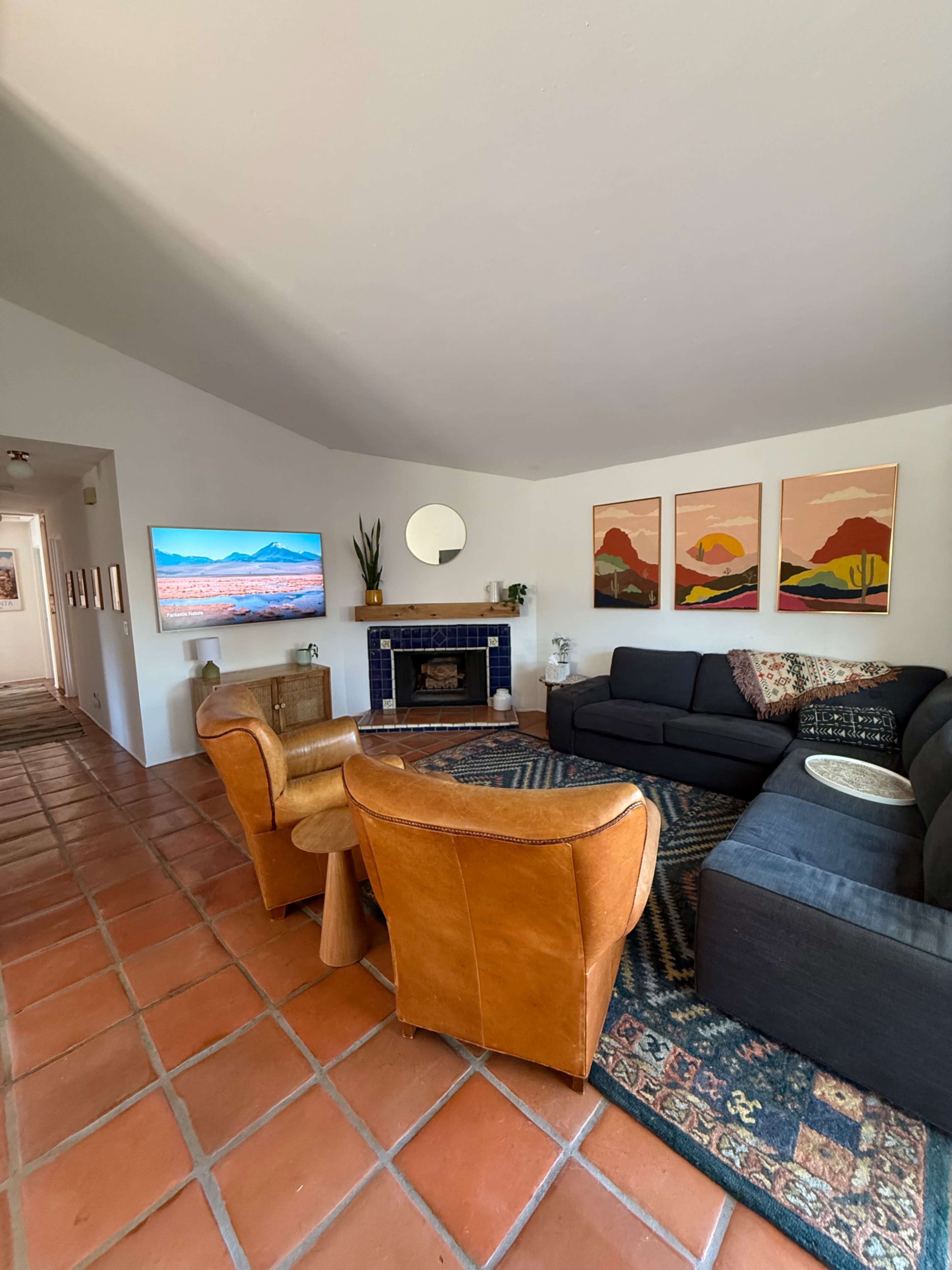 The image shows a living room with a fireplace, two leather armchairs, a dark sectional sofa, and artwork on the walls, all arranged around a patterned area rug.