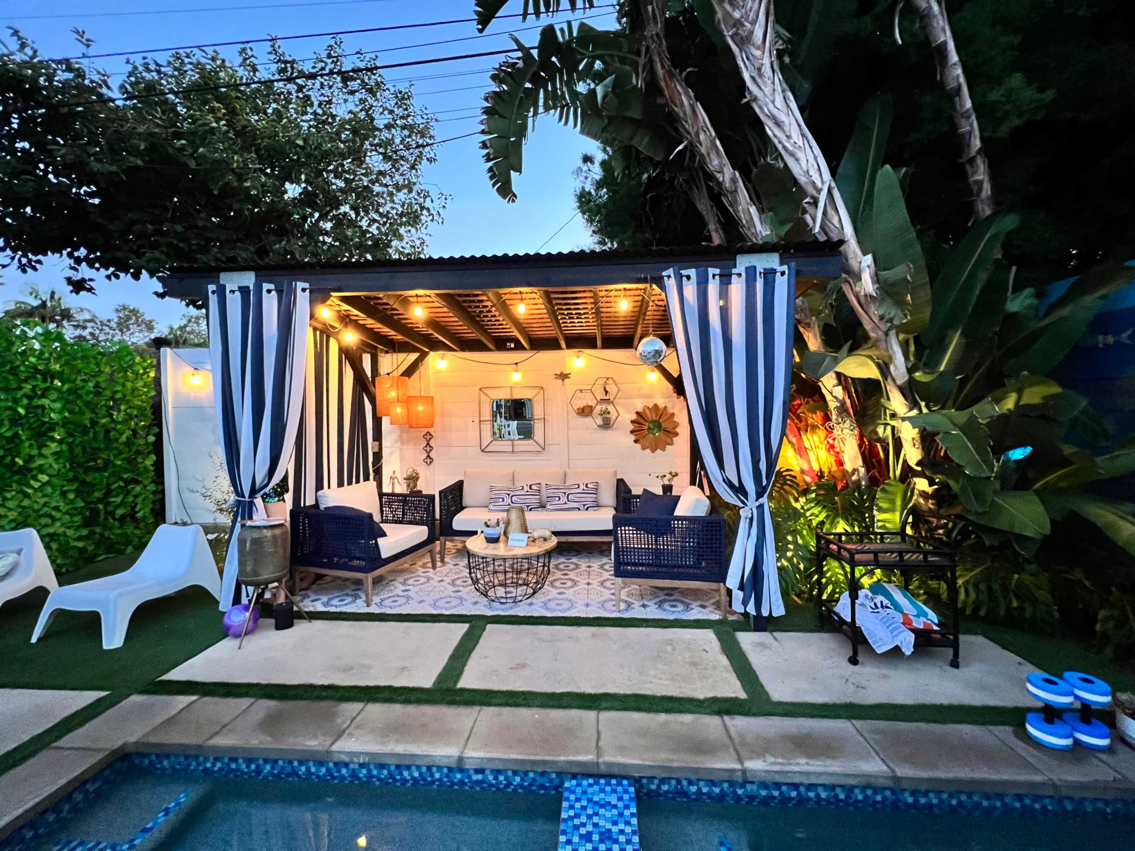 A covered outdoor lounge area with seating and decorative lights is adjacent to a swimming pool.