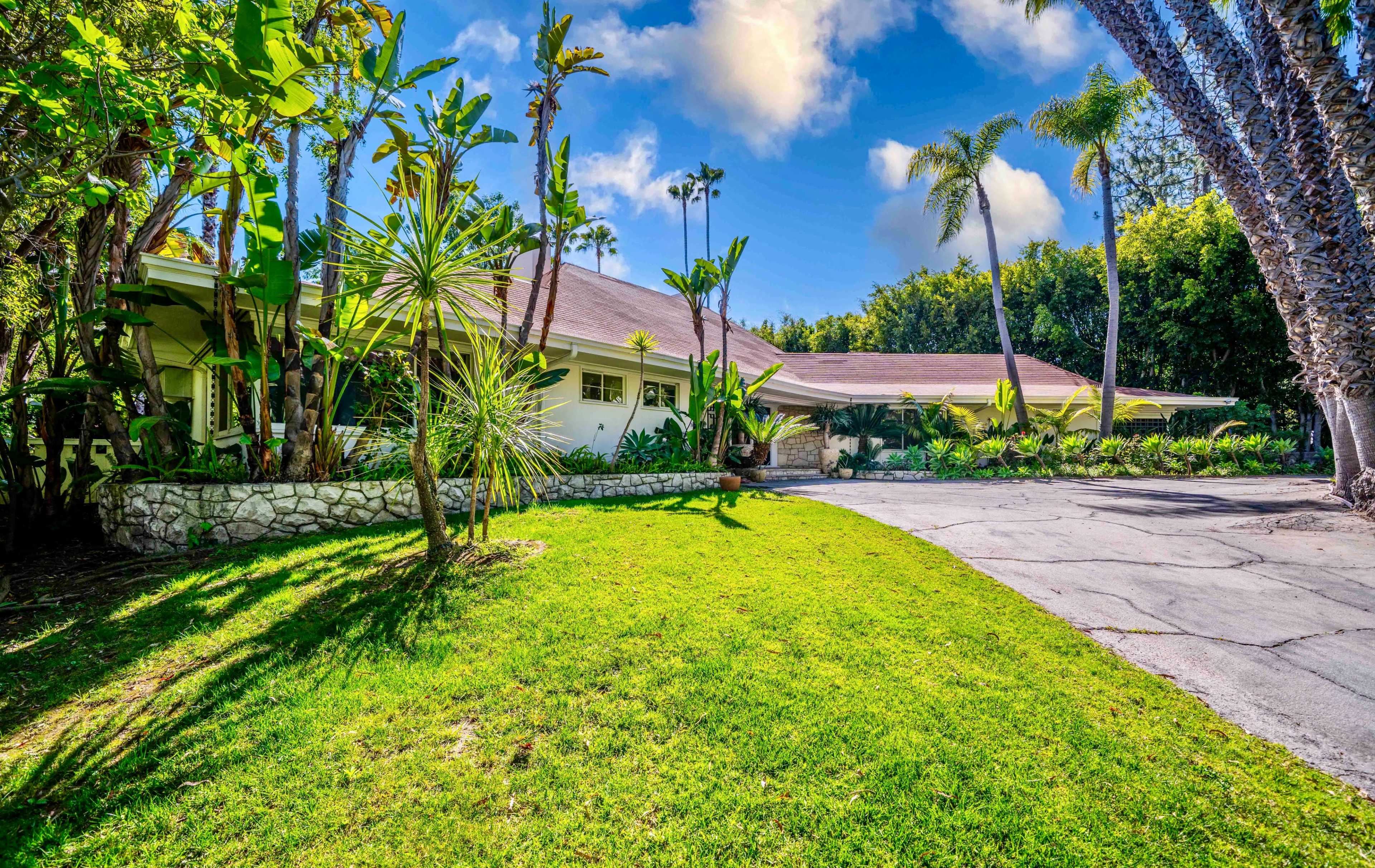 A single-story house with a landscaped yard and palm trees is situated at the end of a paved driveway.