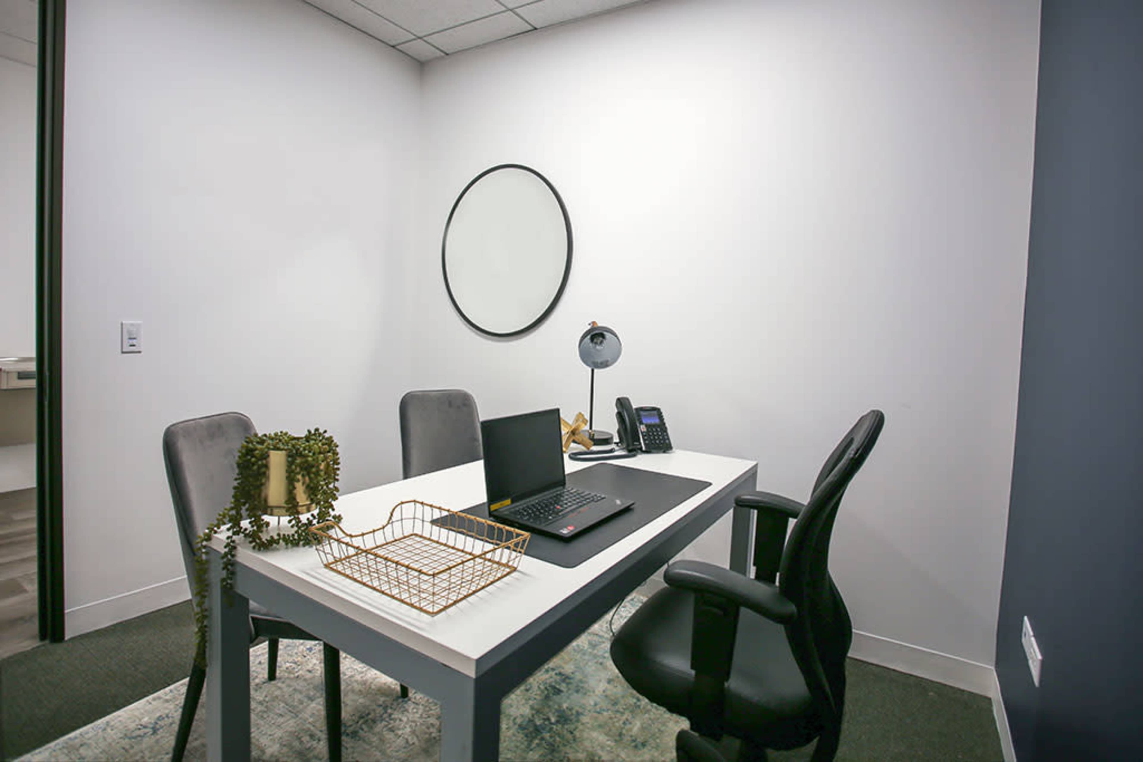 The image shows a minimalist office space featuring a white table, two chairs, a laptop, a desk lamp, and a wall-mounted mirror.
