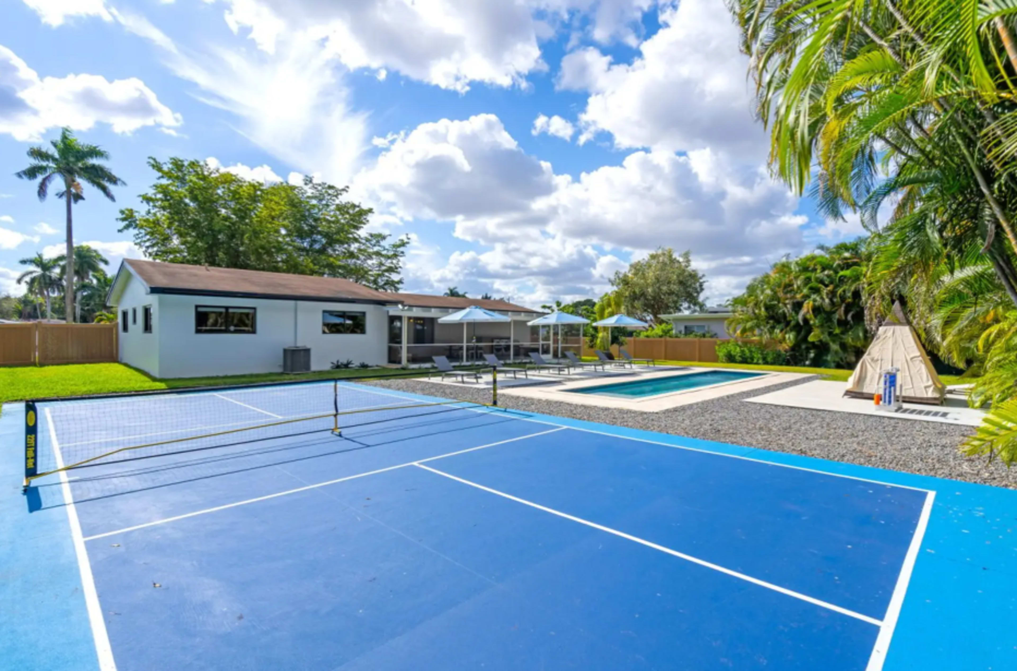 The image shows a residential backyard featuring a blue tennis court, a swimming pool, lounge chairs under umbrellas, and a teepee structure surrounded by greenery.