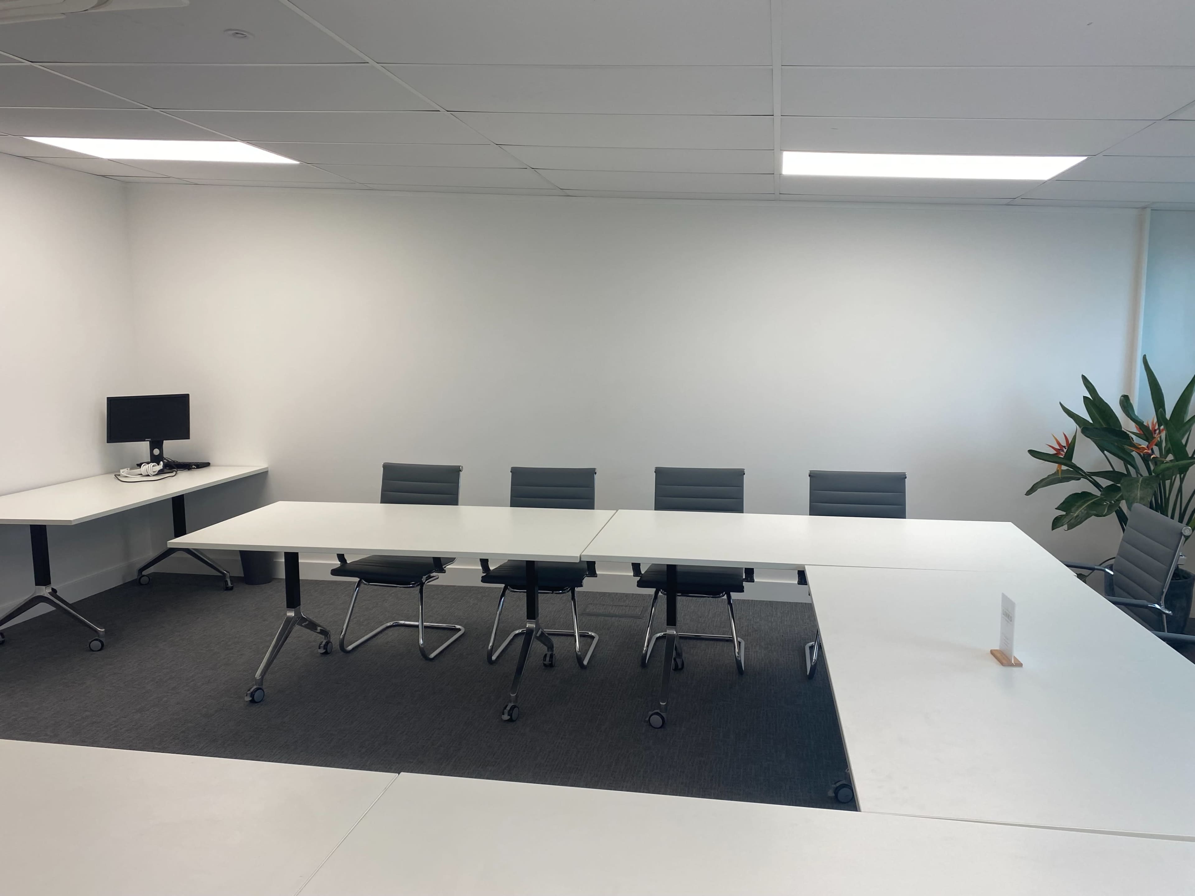 A conference room features a large white table, six gray chairs, a computer monitor on the side, and a potted plant in the corner.