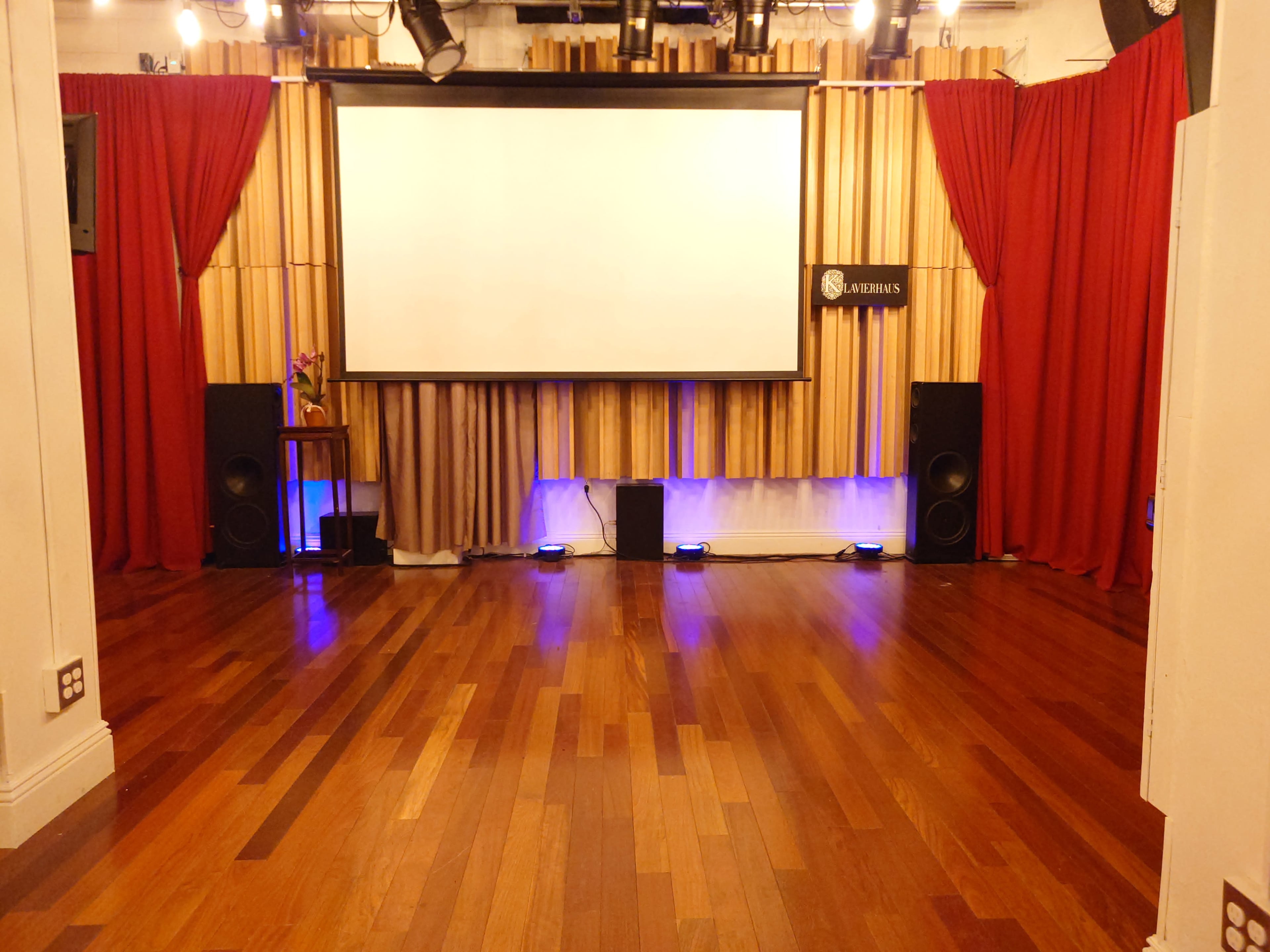 The image shows a spacious event room with wooden floors, a large white screen at the front, and red curtains framing the sides.