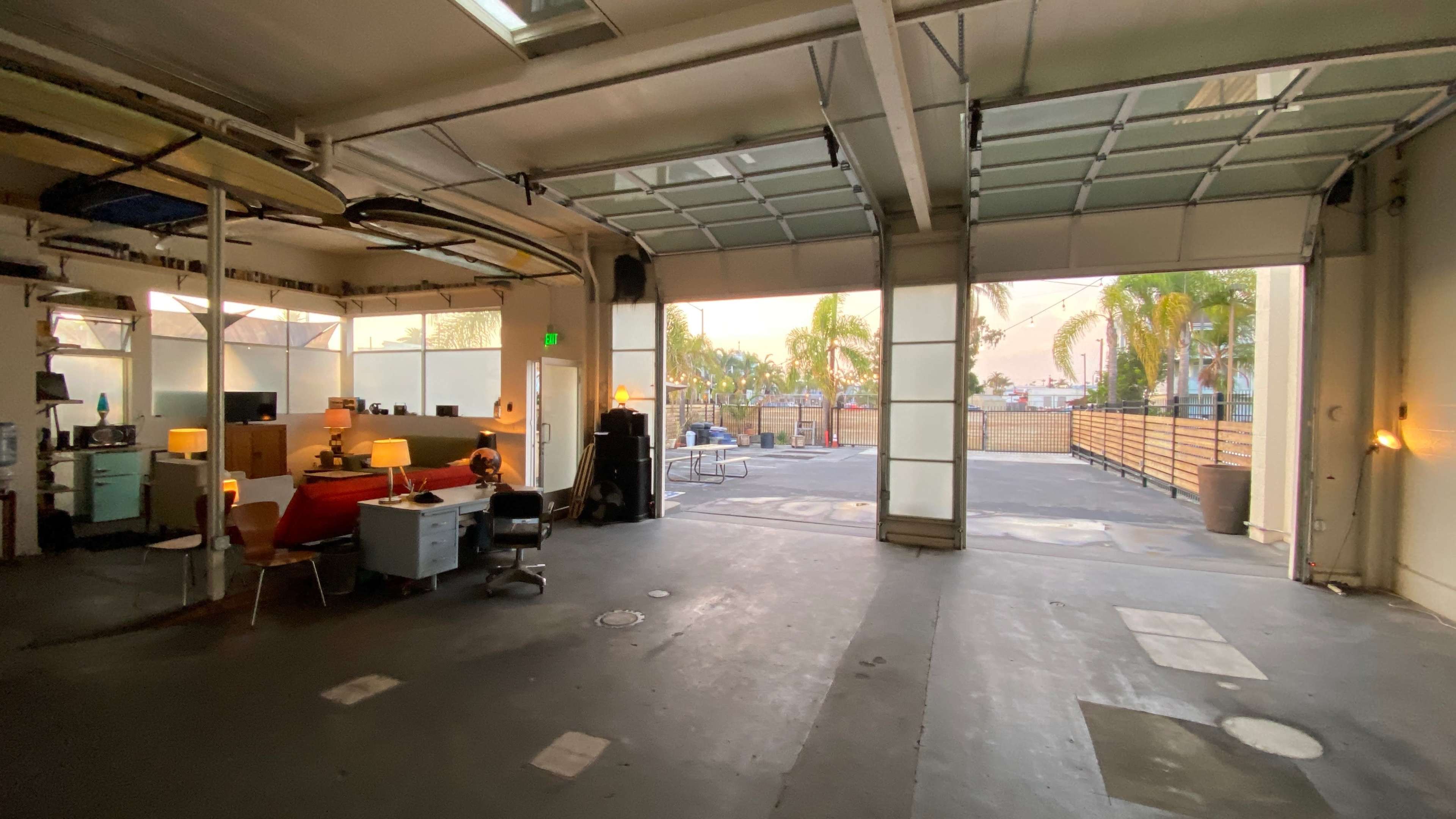 An open garage space features a few office setups with desks and chairs, leading out to an outdoor area with palm trees and a view of a ferris wheel.