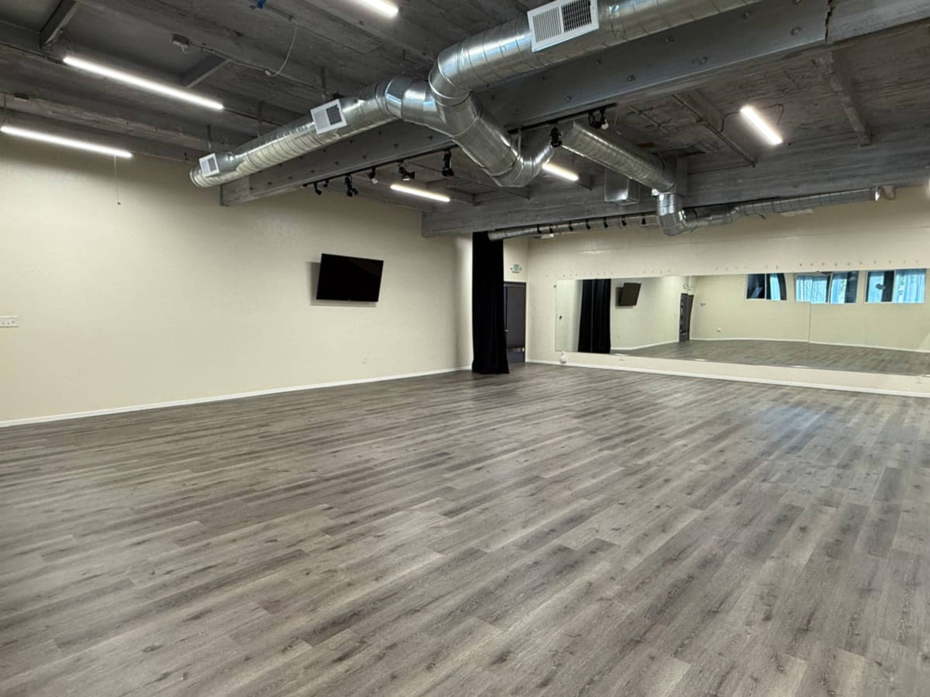 The image shows an empty dance studio with wooden flooring, a large wall mirror, and exposed ductwork along the ceiling.