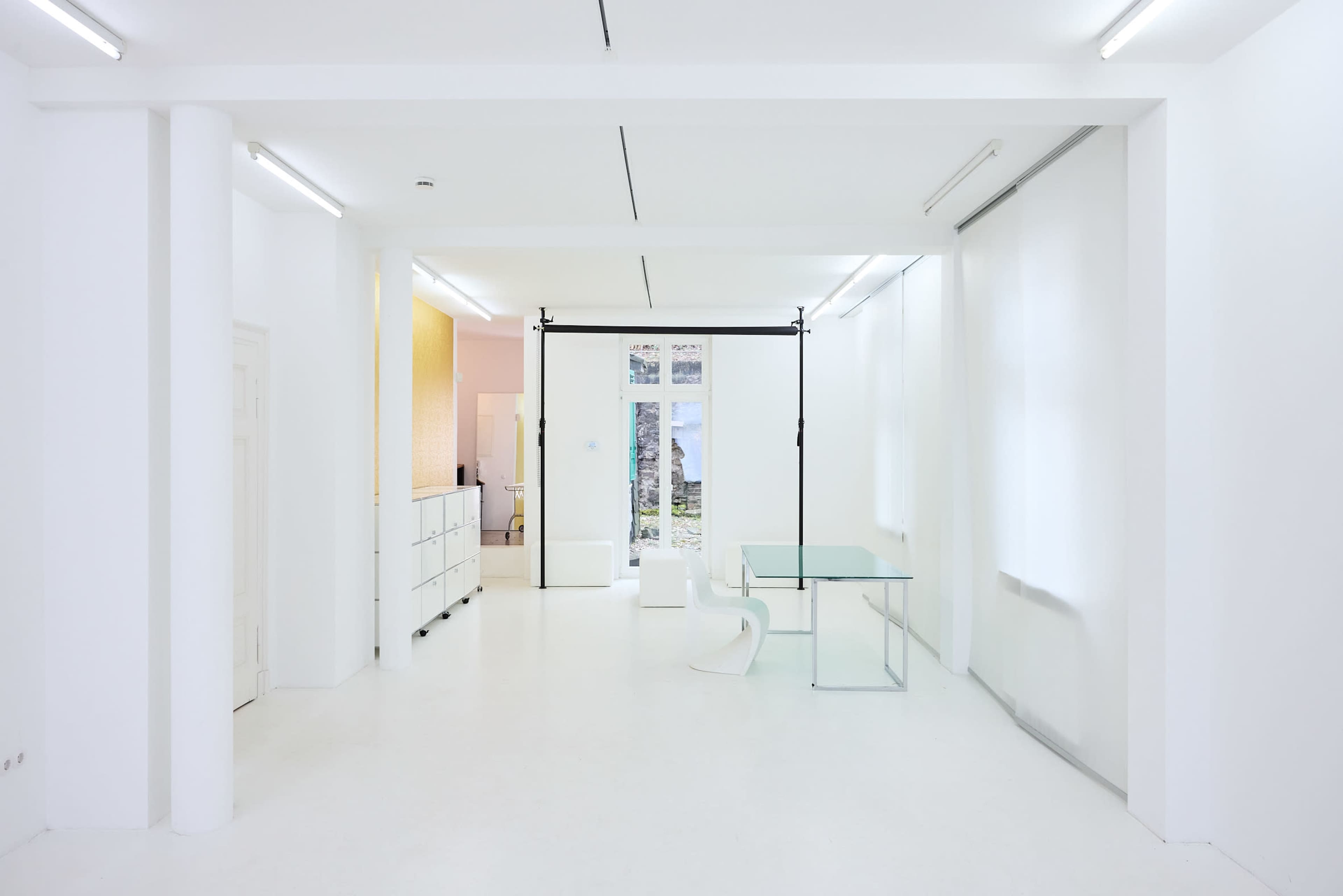 The image shows a spacious, minimalistic interior with white walls, a glass table, and a sliding door leading to an outdoor area.