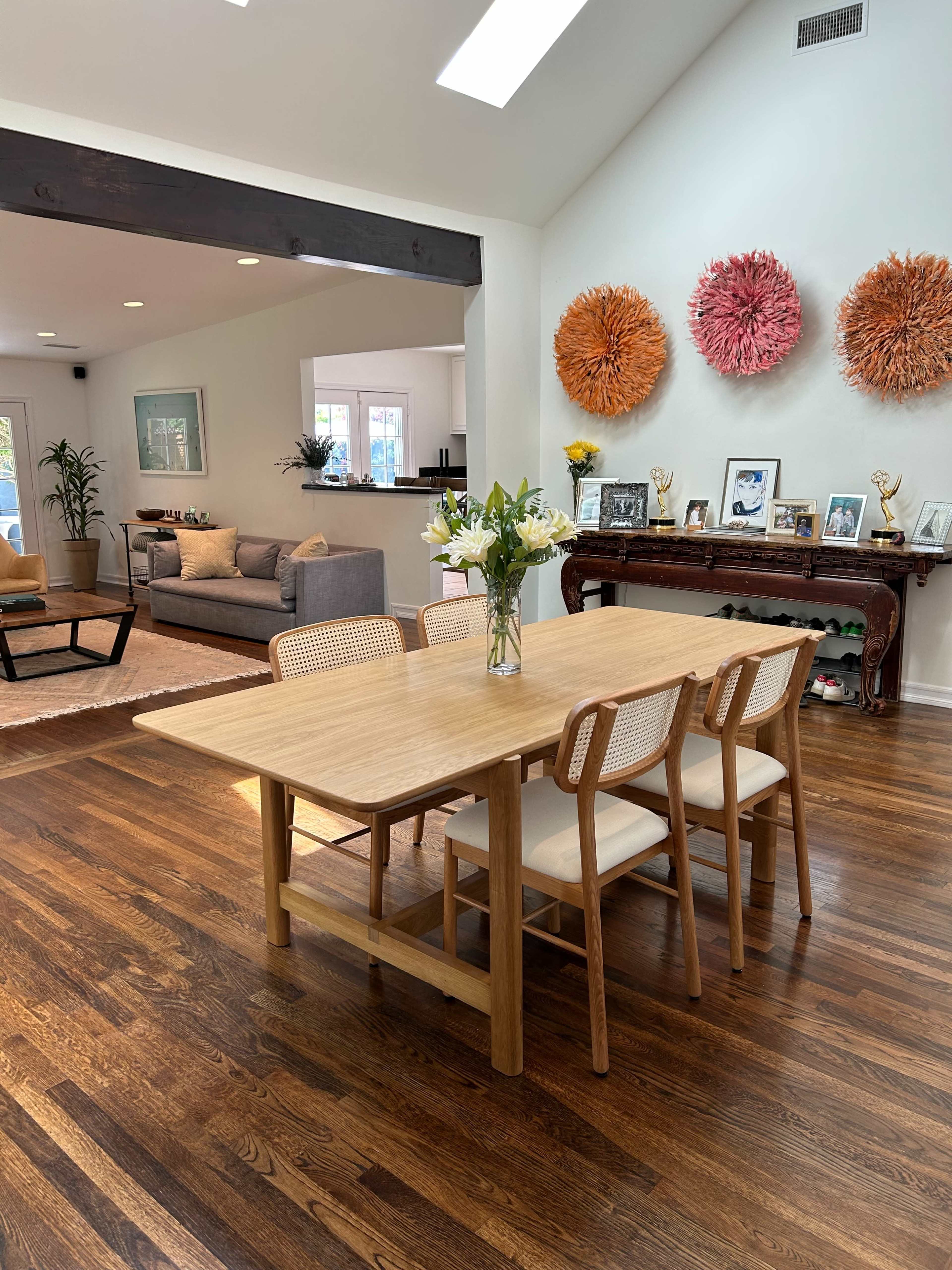 A wooden dining table with five chairs is set in a bright, open-concept living area featuring a gray sofa and decorative wall hangings.