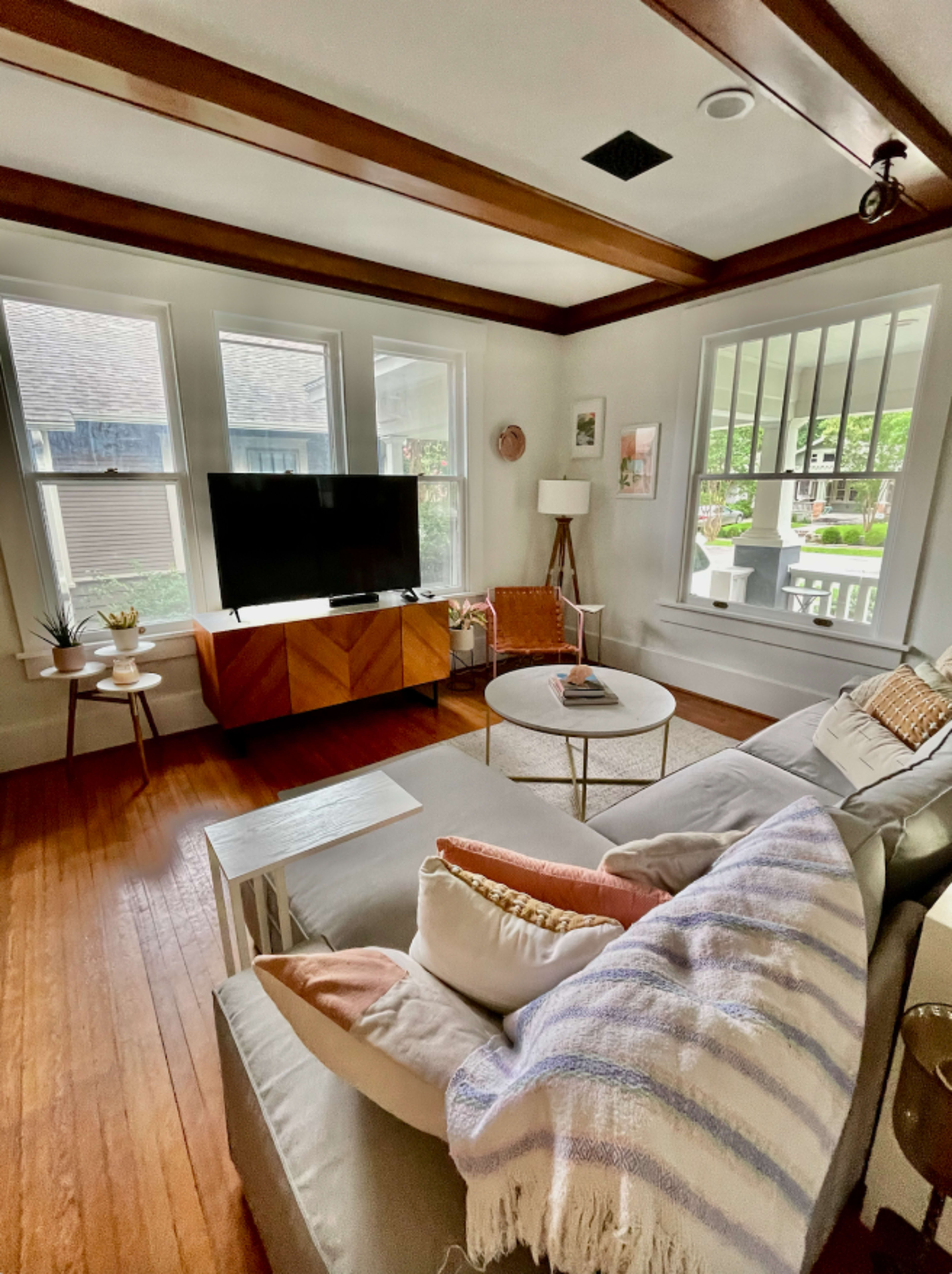 The living room features a sectional sofa with decorative pillows, a coffee table, a wooden media cabinet holding a television, and large windows providing natural light.