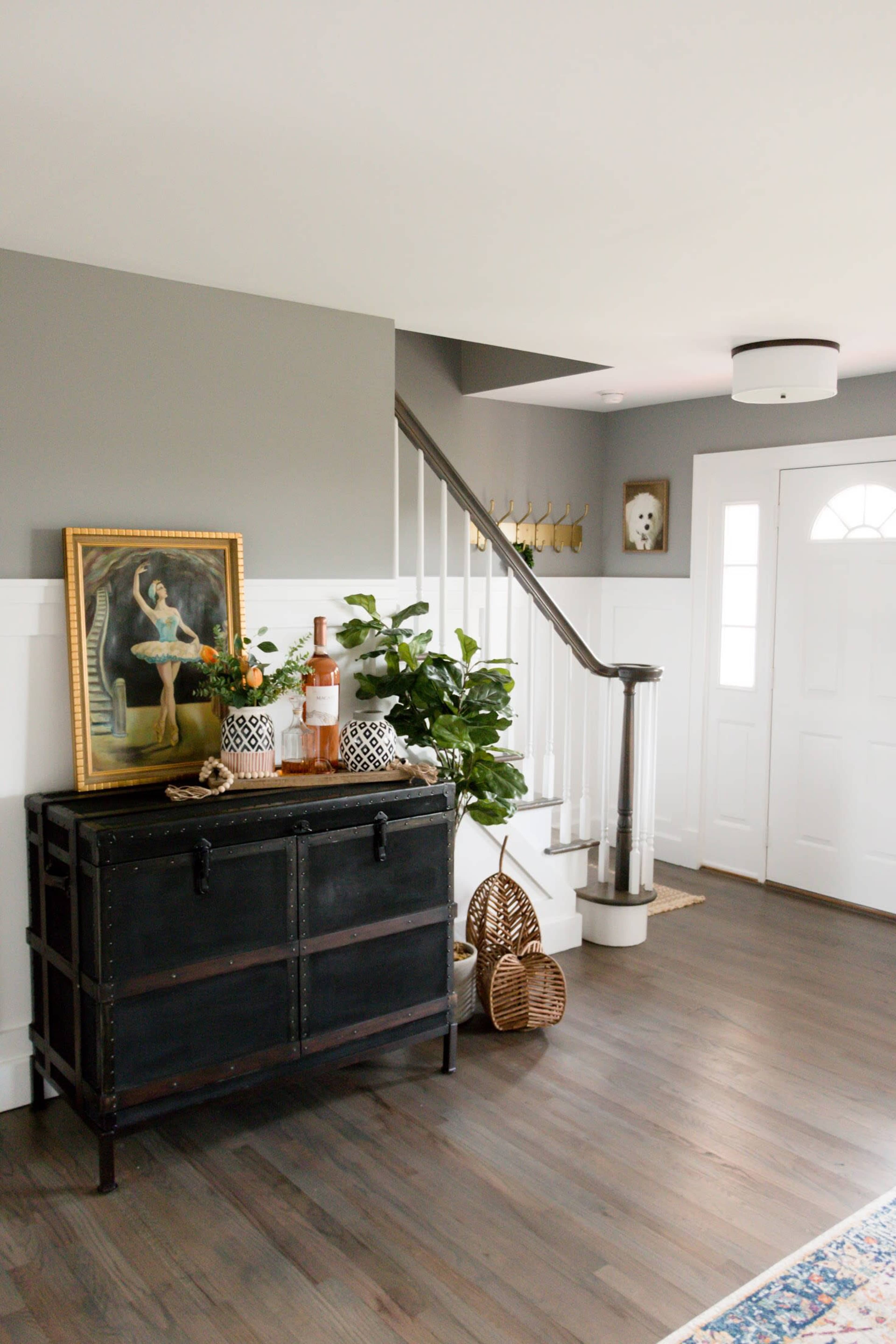 An indoor entryway with a staircase, featuring a black storage chest, a framed painting, plants, and decorative items near a front door.