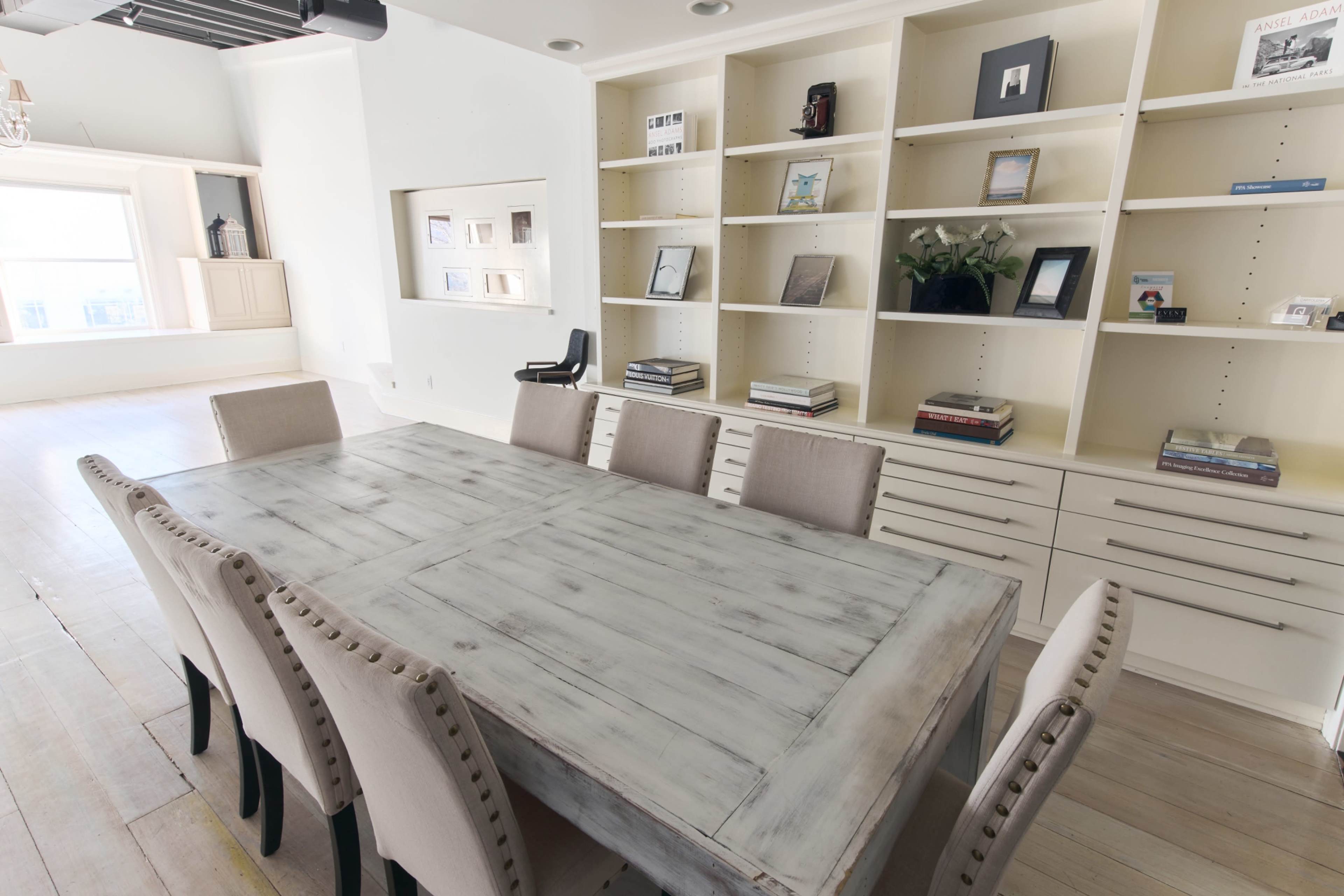 A dining area features a long wooden table surrounded by beige upholstered chairs, with a built-in bookshelf displaying various items on the wall.