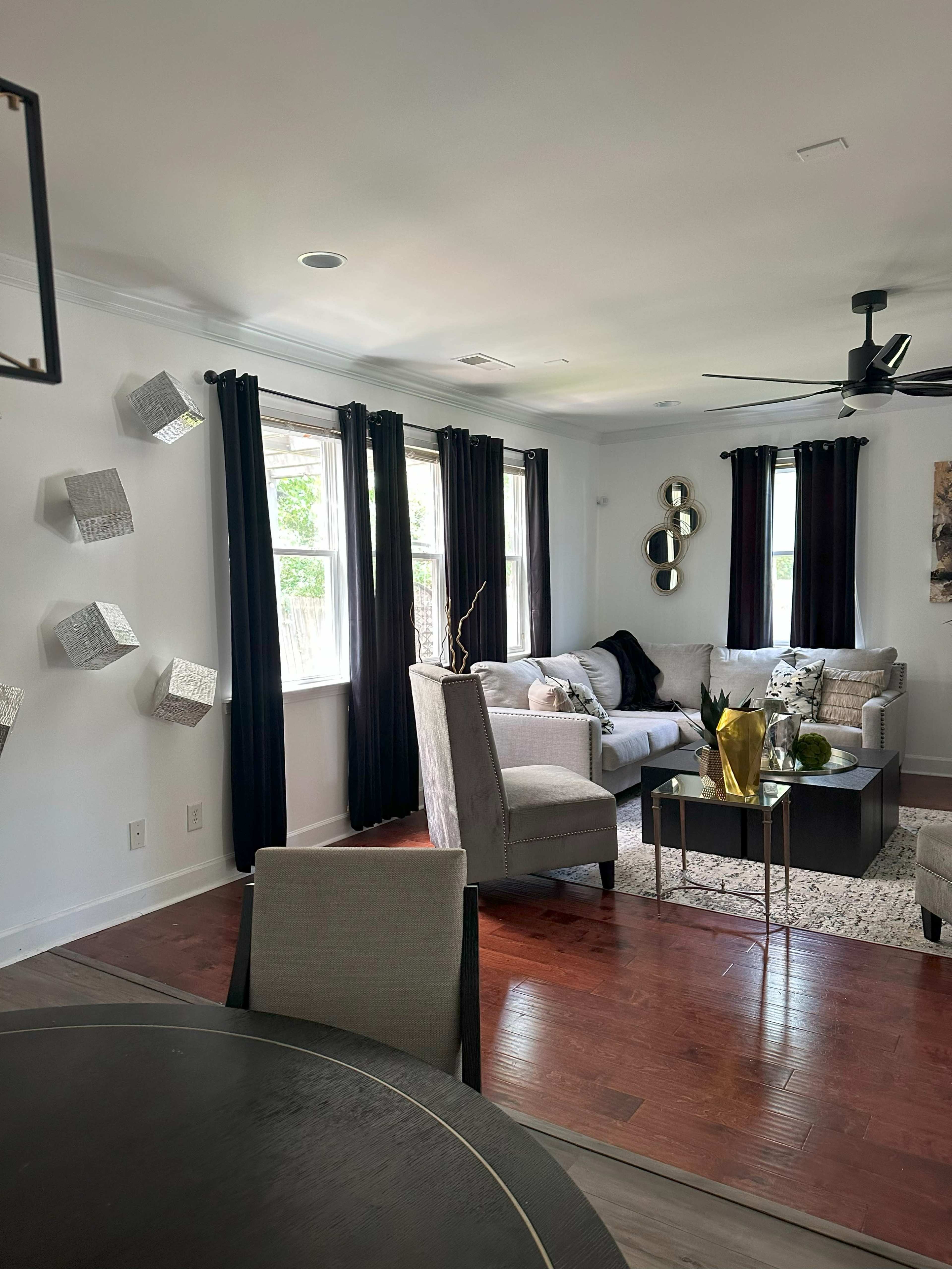 A modern living room features a light-colored sofa, dark curtains, and decorative wall accents, with a coffee table and seating area arranged around it.
