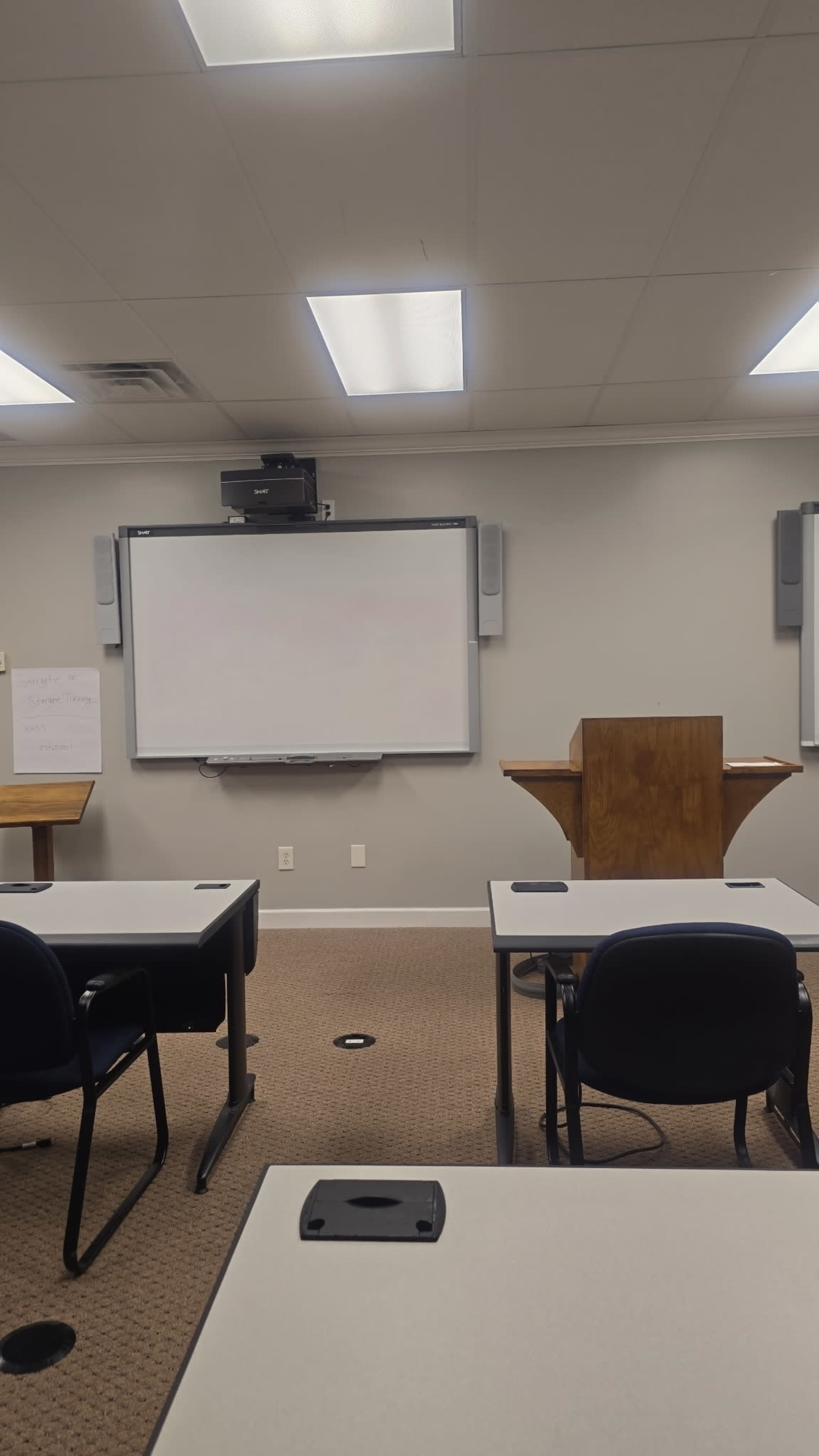 The image shows an empty classroom with several tables and chairs, a large whiteboard at the front, and a wooden lectern on one side.