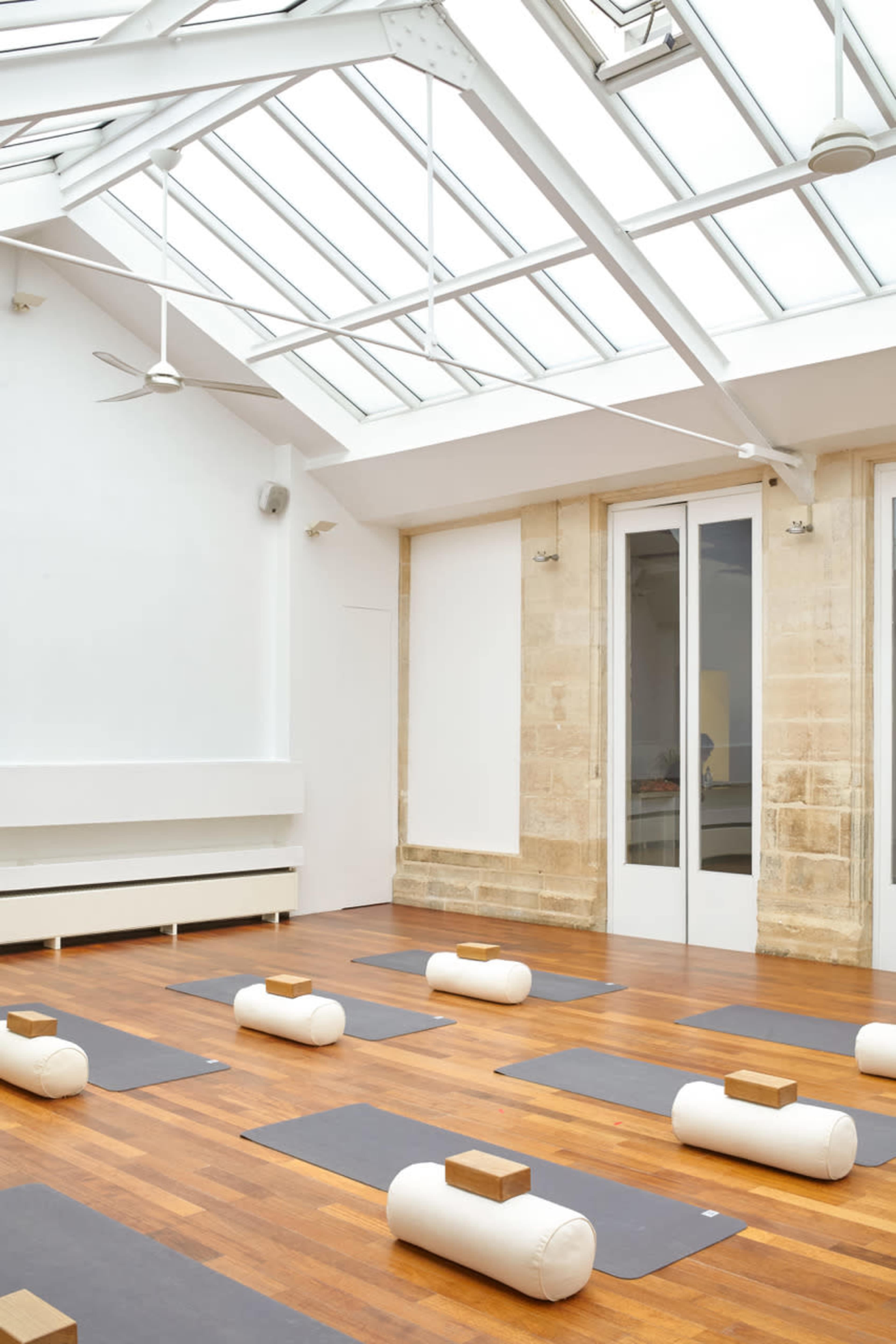 A bright, minimalist yoga studio with wooden floors, several yoga mats arranged on the ground, and bolsters with blocks placed on top.