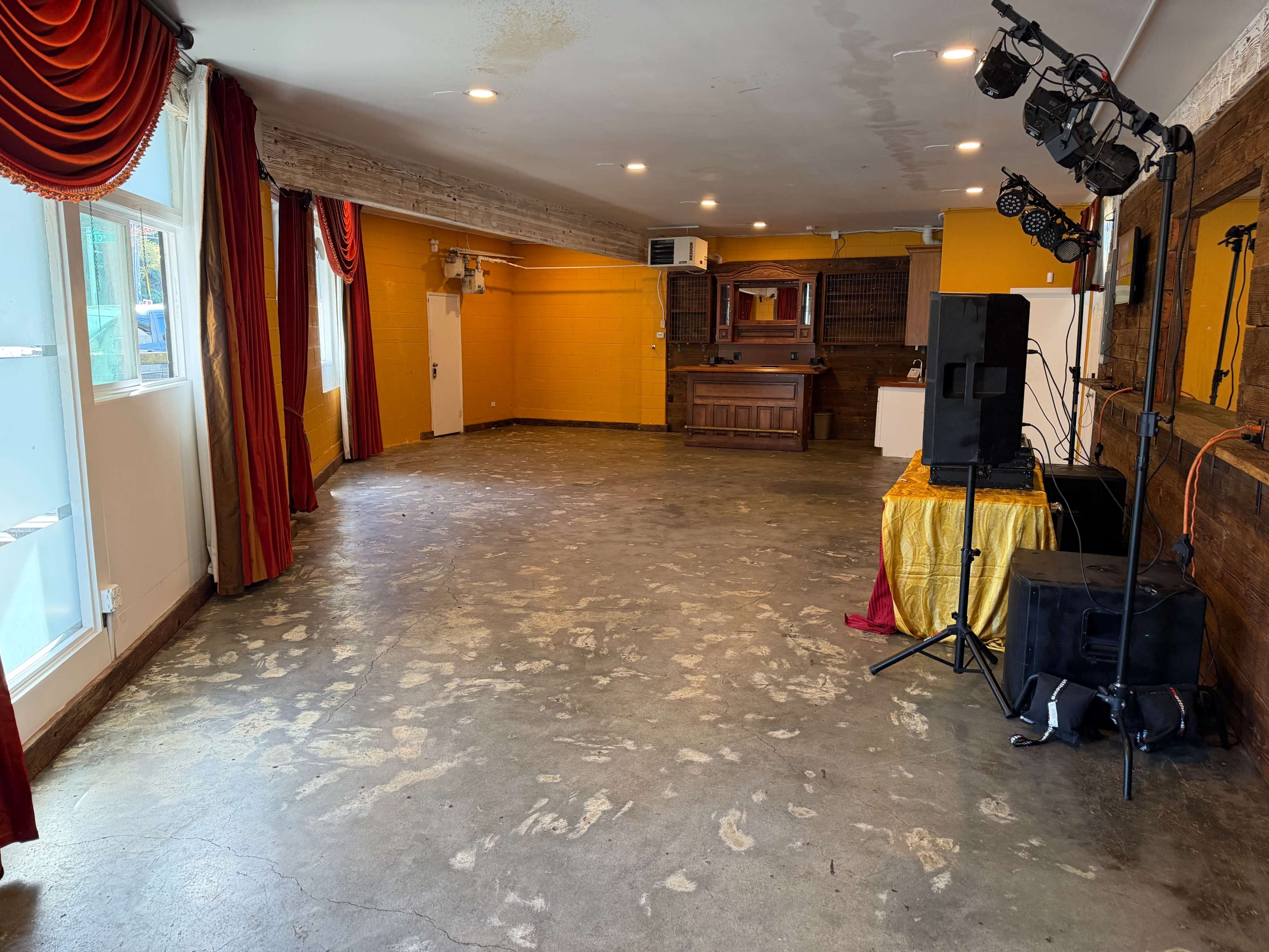 The room features a spacious, empty area with a partially polished concrete floor, orange walls, and a stage at one end equipped with sound equipment.