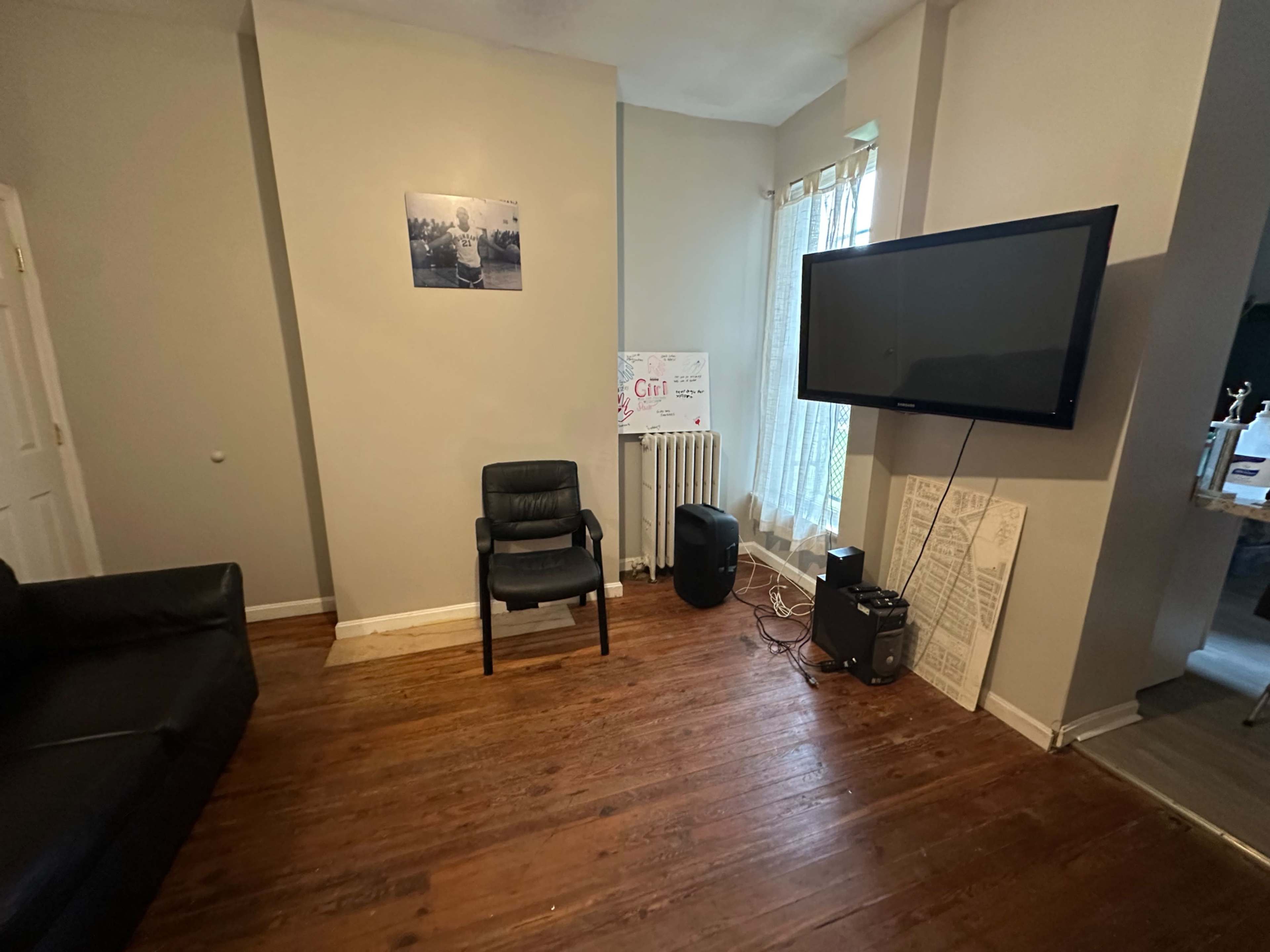 The image shows a living room with a black chair, a mounted flat-screen TV, and wooden flooring, along with a radiator and a whiteboard.