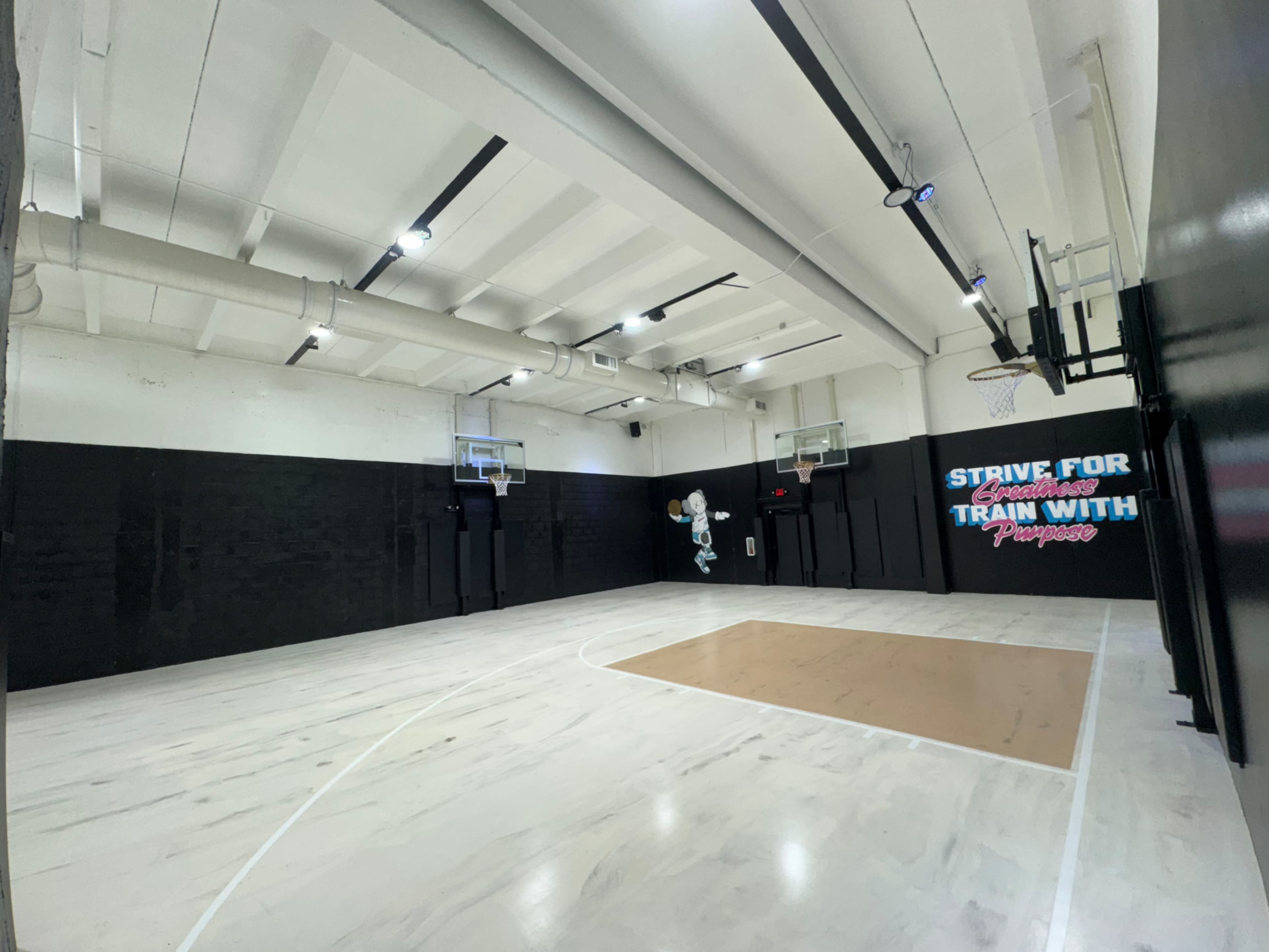 The image shows an indoor basketball court with two hoops, a smooth wooden floor, and a motivational quote on the wall.