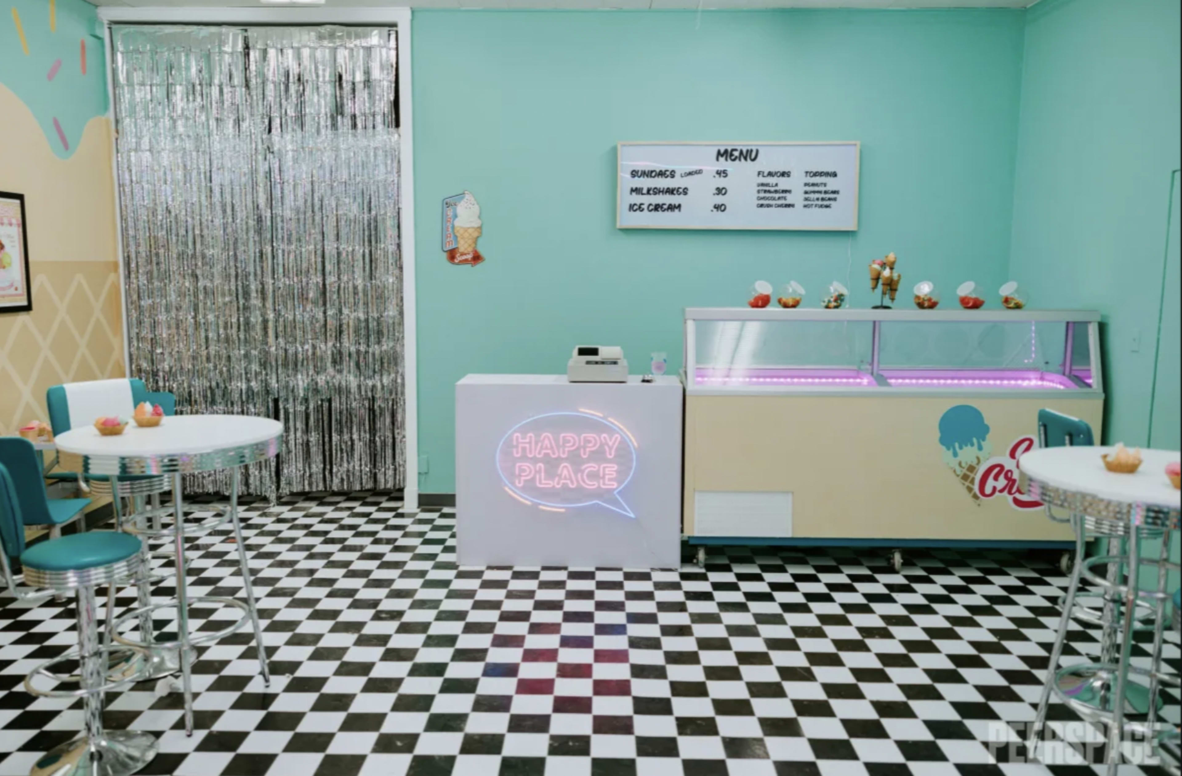 A colorful ice cream shop with a checkered floor, featuring a neon "HAPPY PLACE" sign, a service counter, and a menu board on the wall.