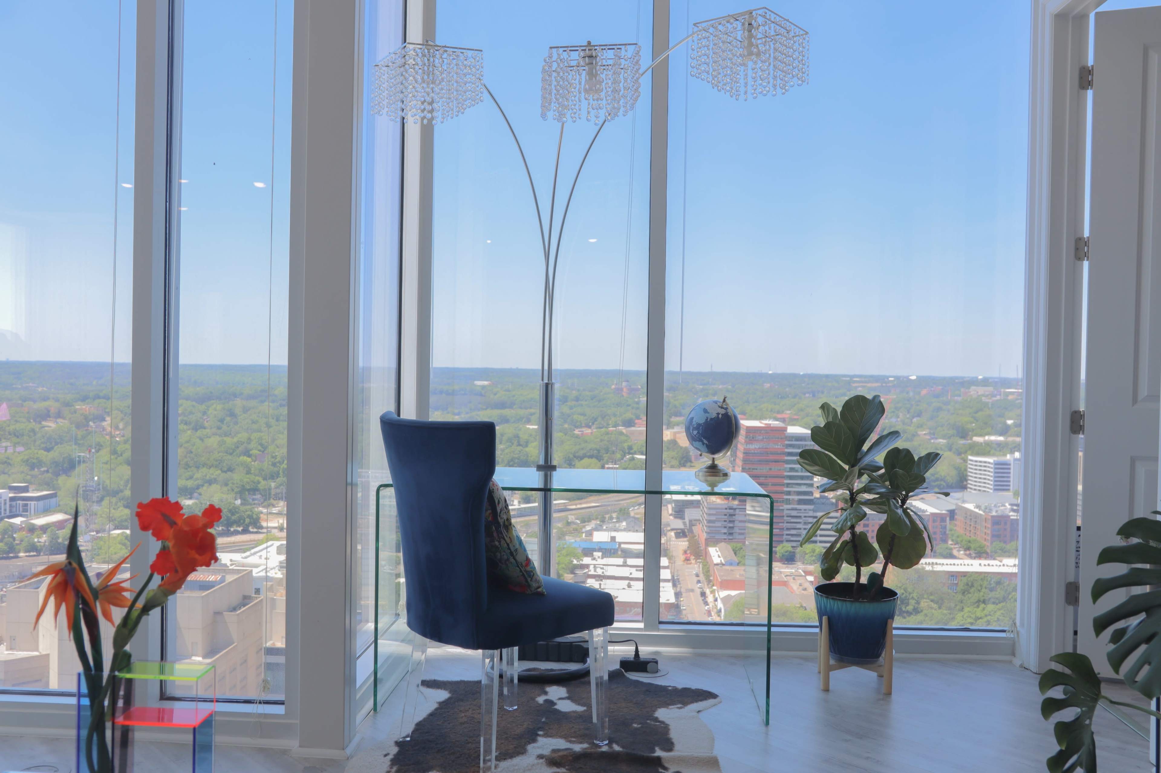 A modern corner office with a clear glass desk, a blue chair, decorative plants, and a large window offering a panoramic view of the city.