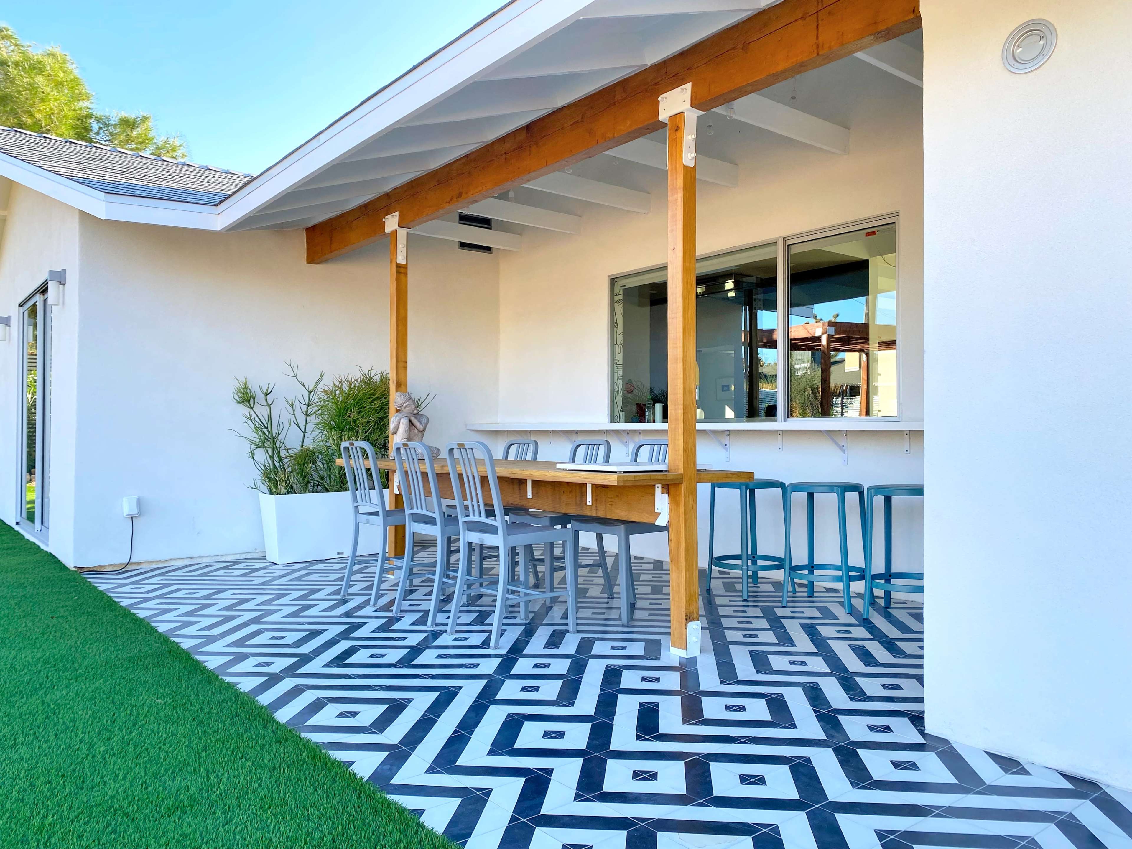 The image shows a patio area with a wooden table and chairs atop a geometric patterned floor, adjacent to a white house with large windows.