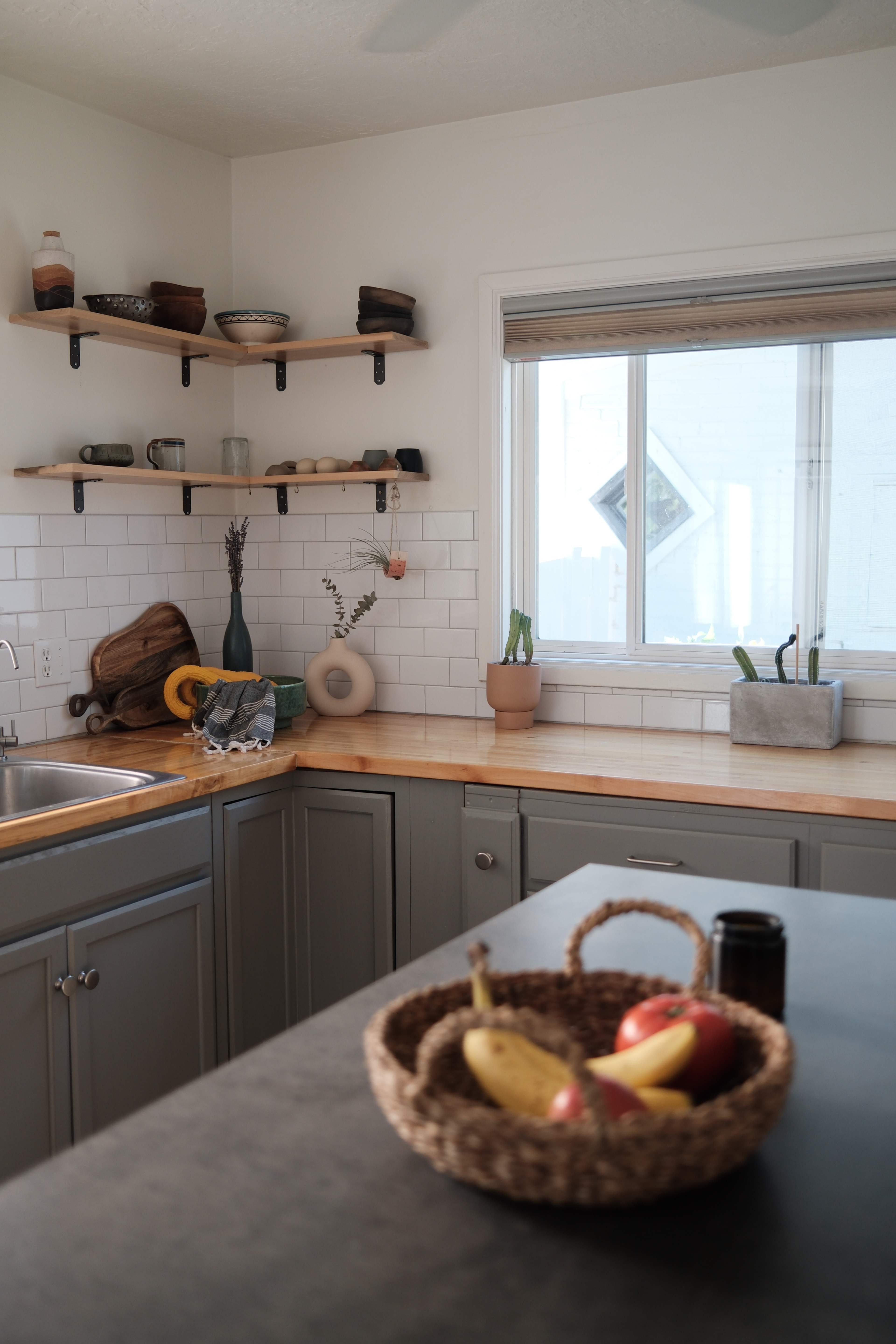 A modern kitchen features wooden shelves with various pottery items, a stainless steel sink, and a counter holding a woven basket of fruit.