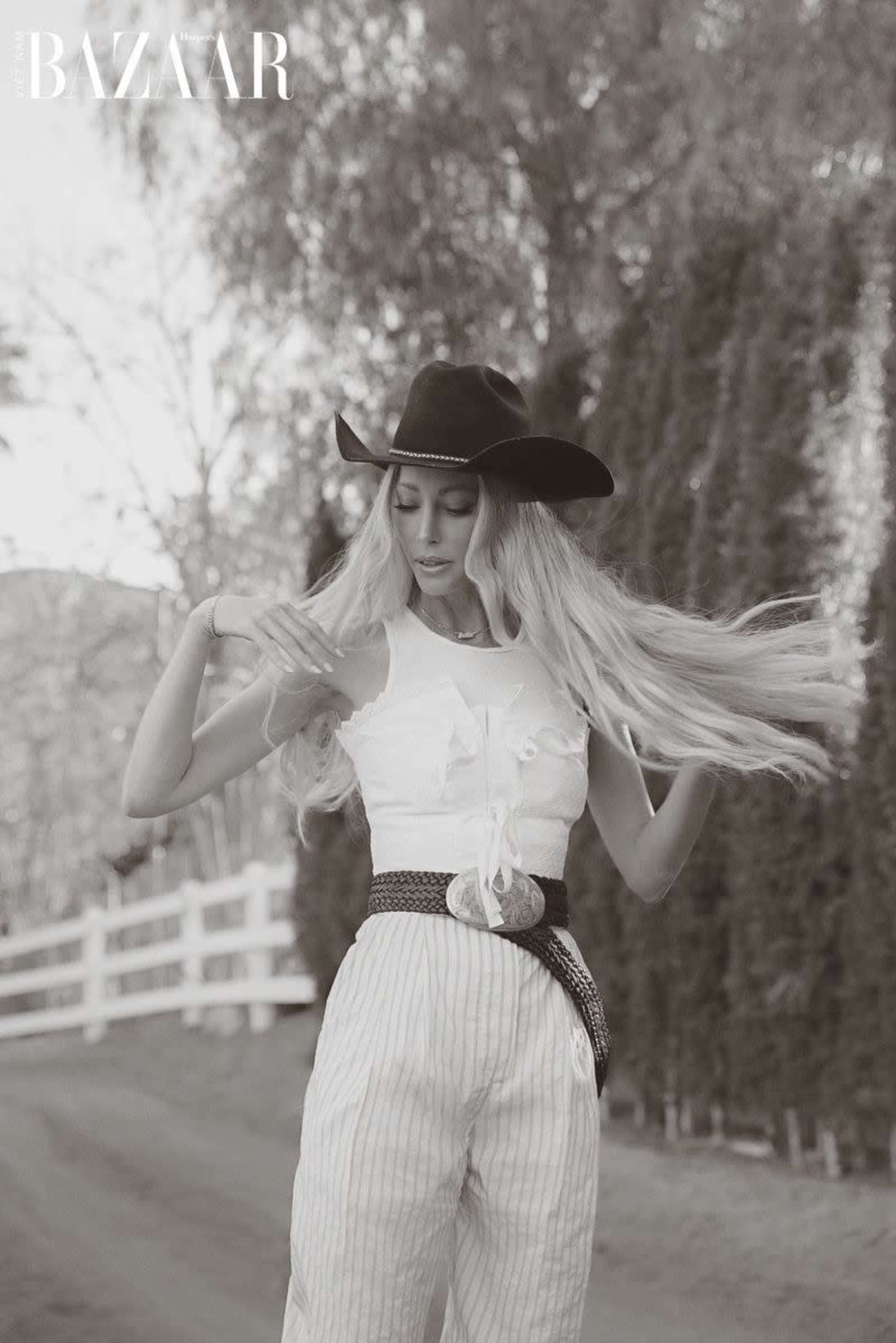 A woman adjusts her hair while wearing a hat and a white top paired with striped pants in an outdoor setting.