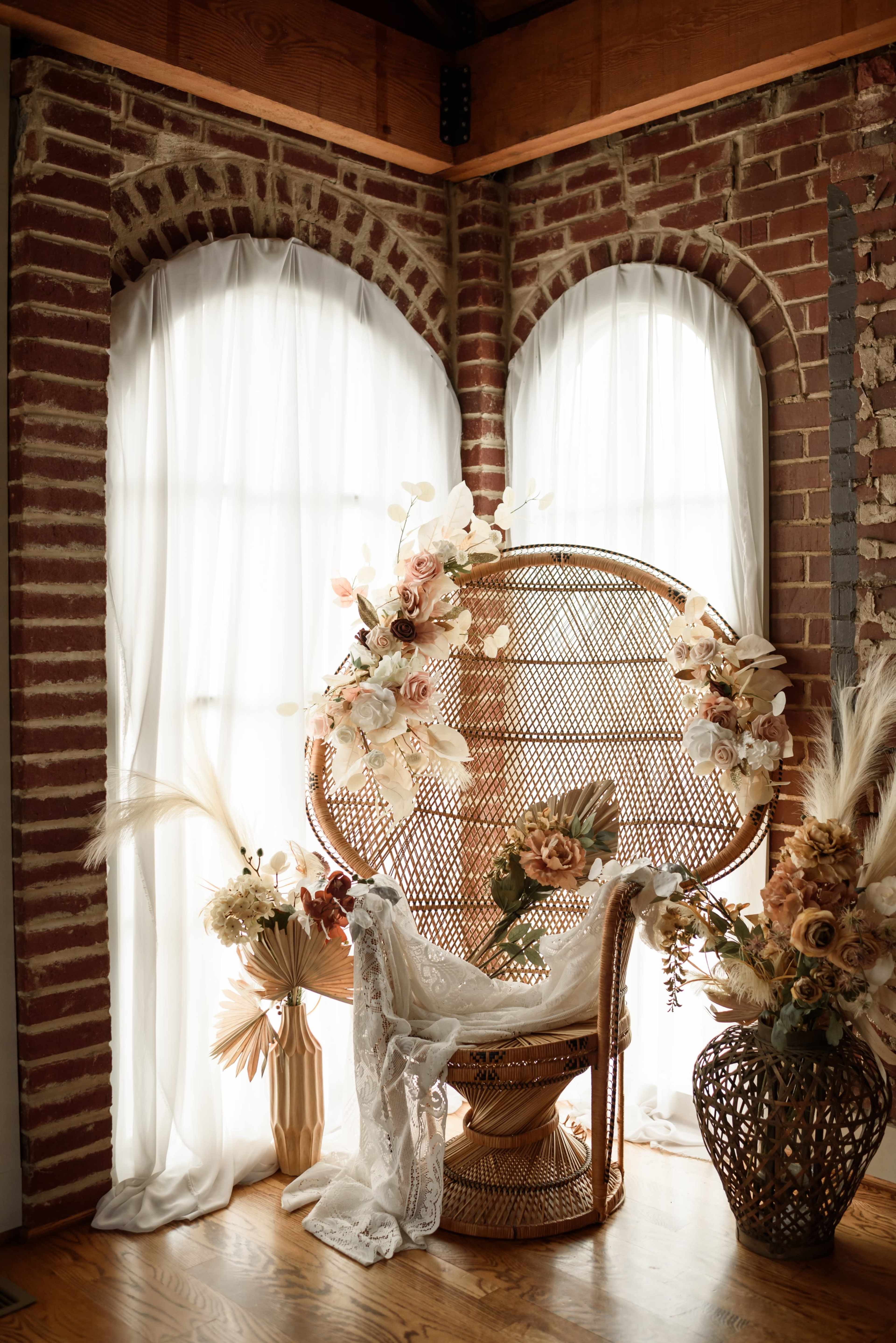 A woven chair adorned with floral arrangements is positioned beside large windows with flowing white curtains in a brick-walled room.