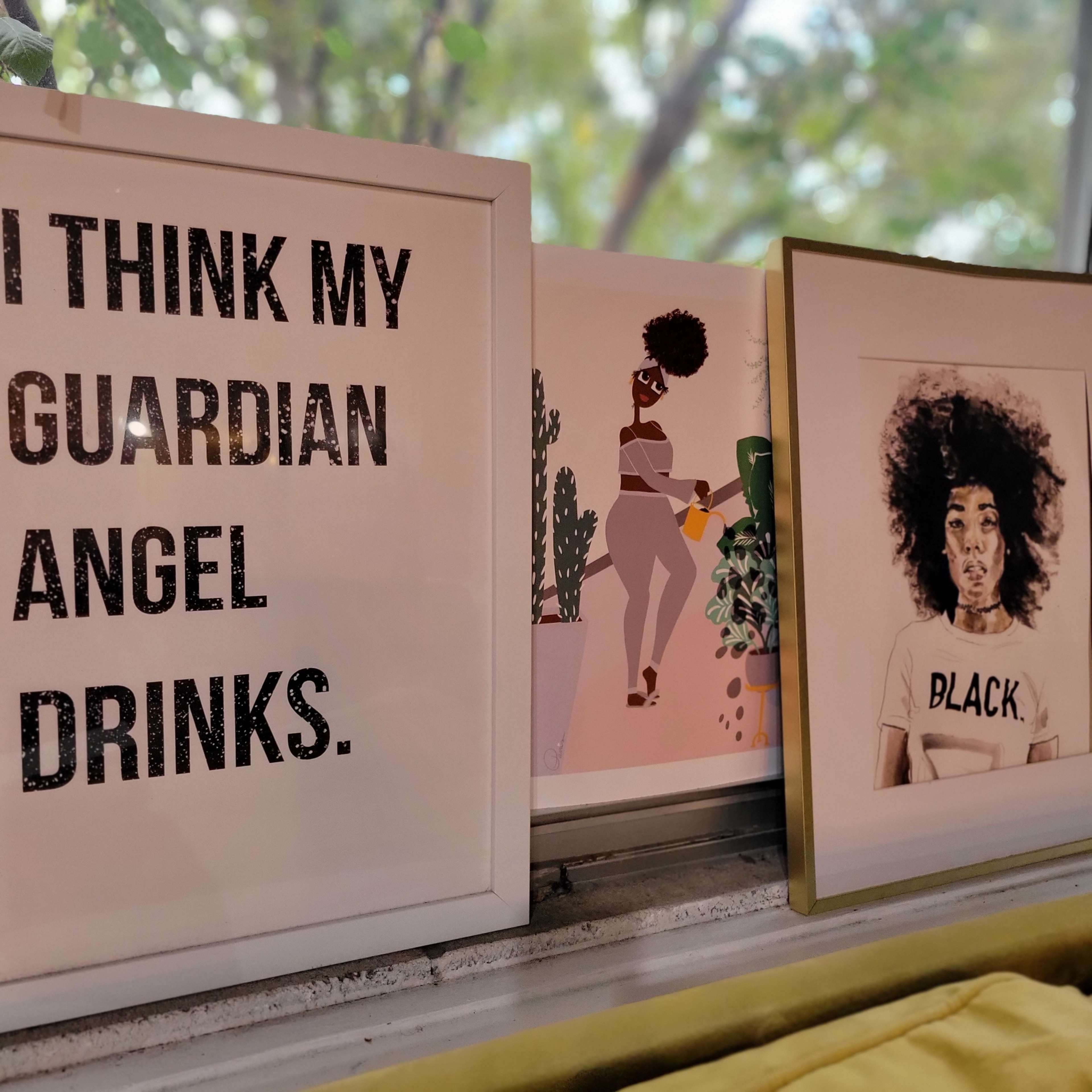 Three framed artworks displayed on a windowsill, including a bold text piece, a colorful illustration of a woman with a plant, and a portrait of a person with an afro.