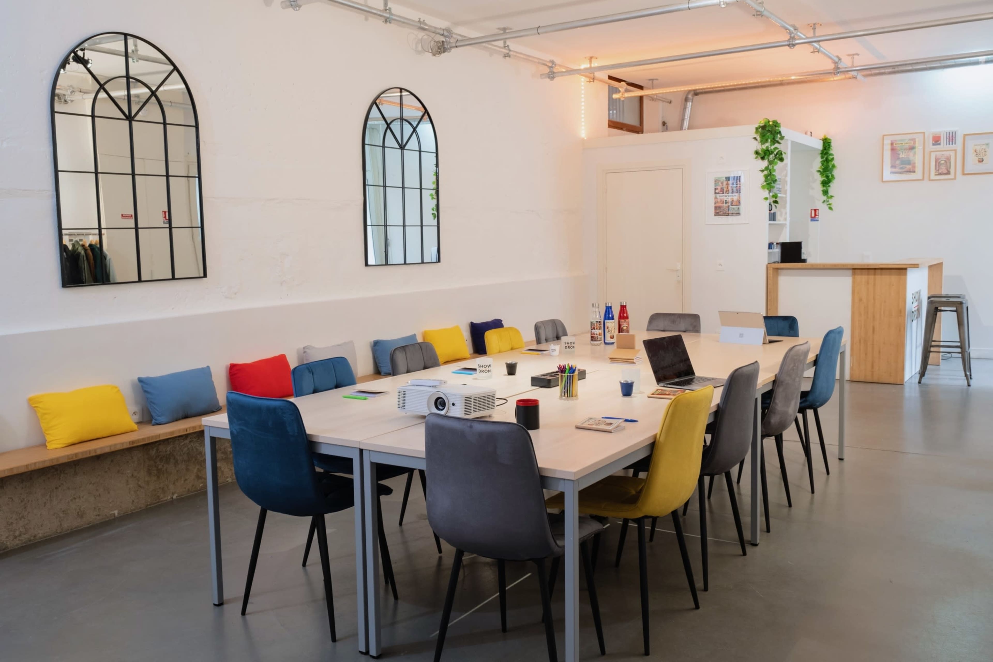 The image depicts a modern meeting room with a long table surrounded by colorful chairs, and a projector set up on one end.