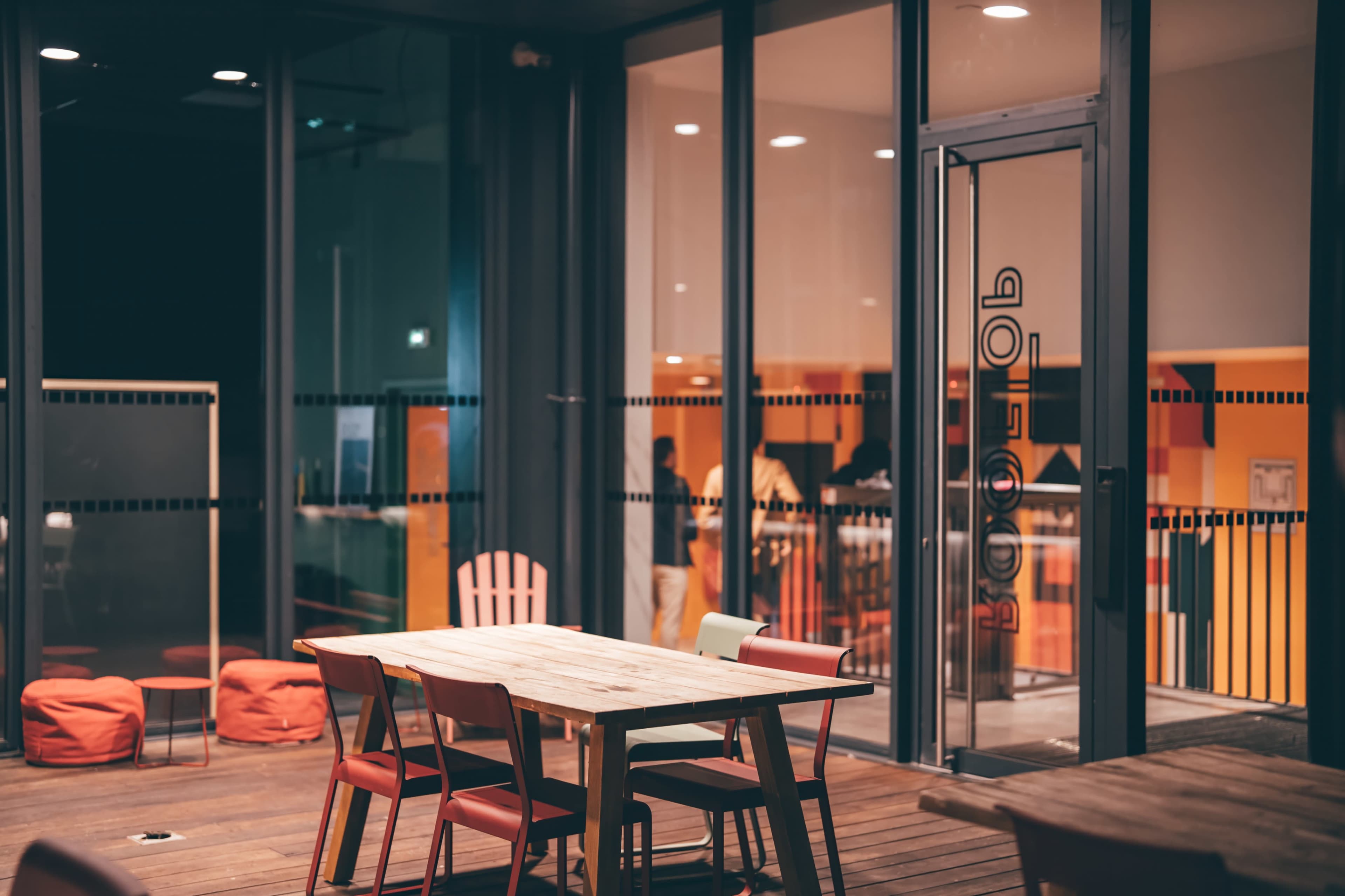 The image shows a modern indoor space with a wooden table and colorful chairs in front of a glass entrance.