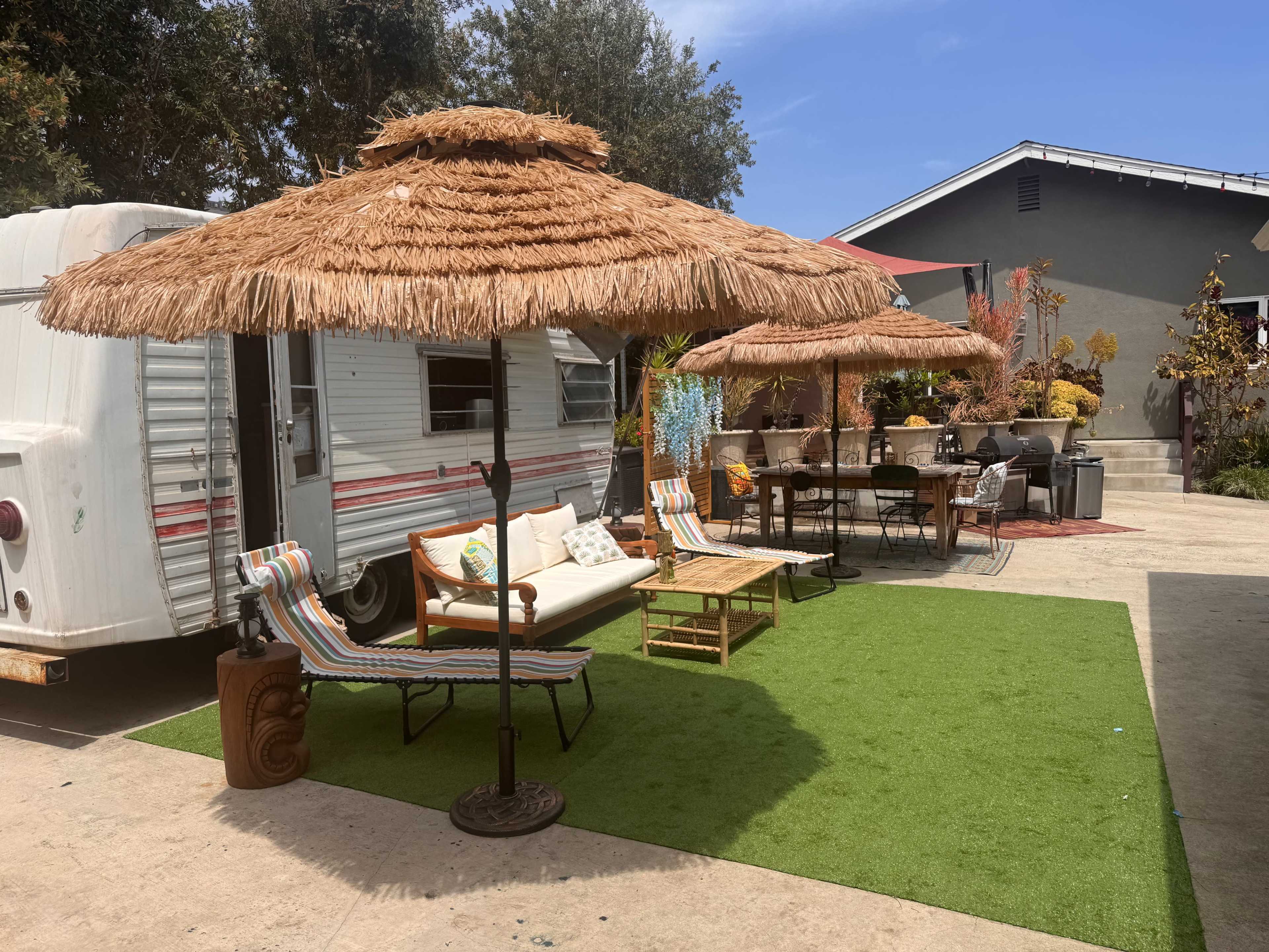 A vintage trailer is parked beside a green turf area with two thatched umbrellas, lounge chairs, and a dining table amidst various potted plants.