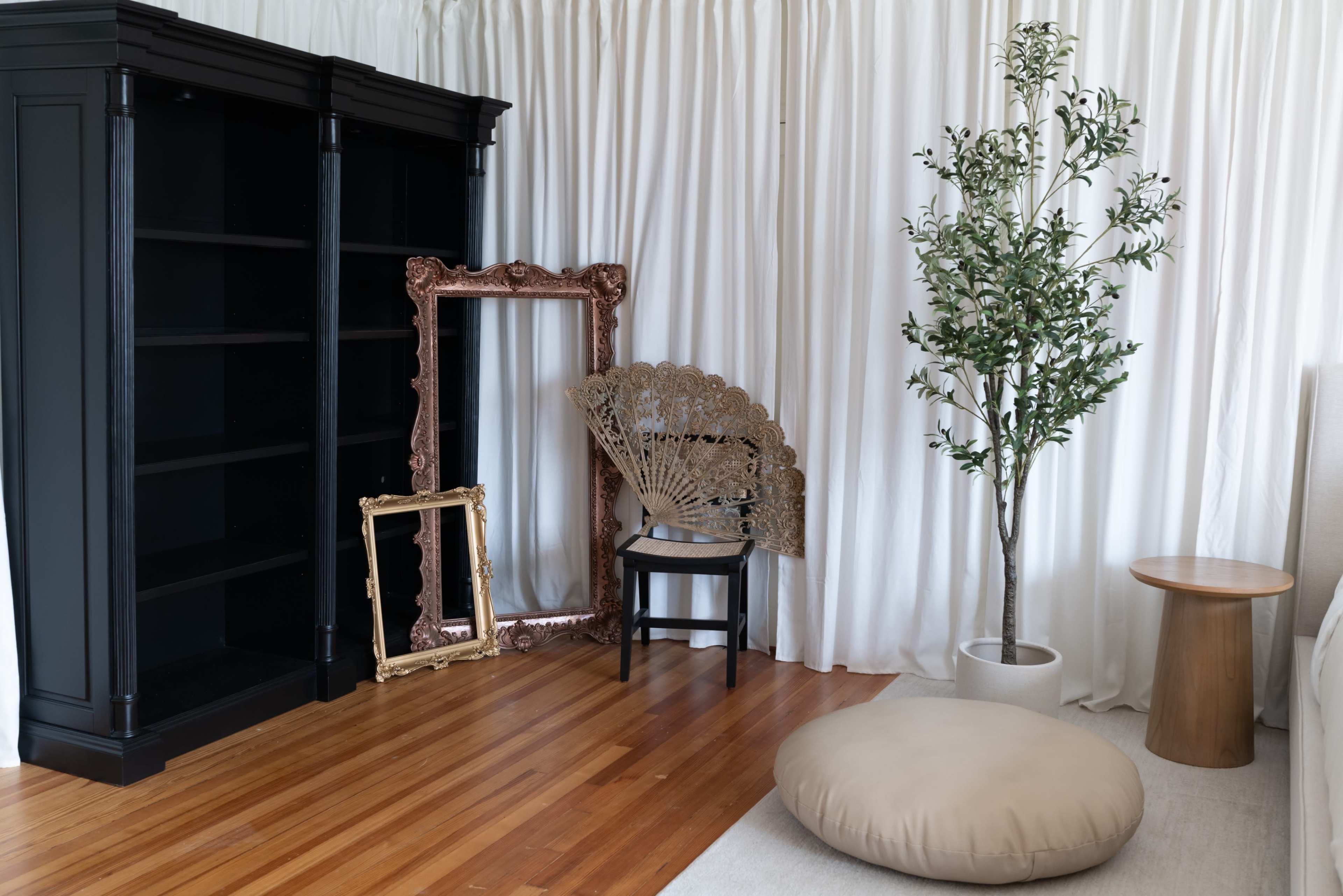 The scene features a minimalist interior with a black bookshelf, a decorative fan, two ornate frames, a potted plant, a round stool, and a large floor cushion, all set against white curtains.