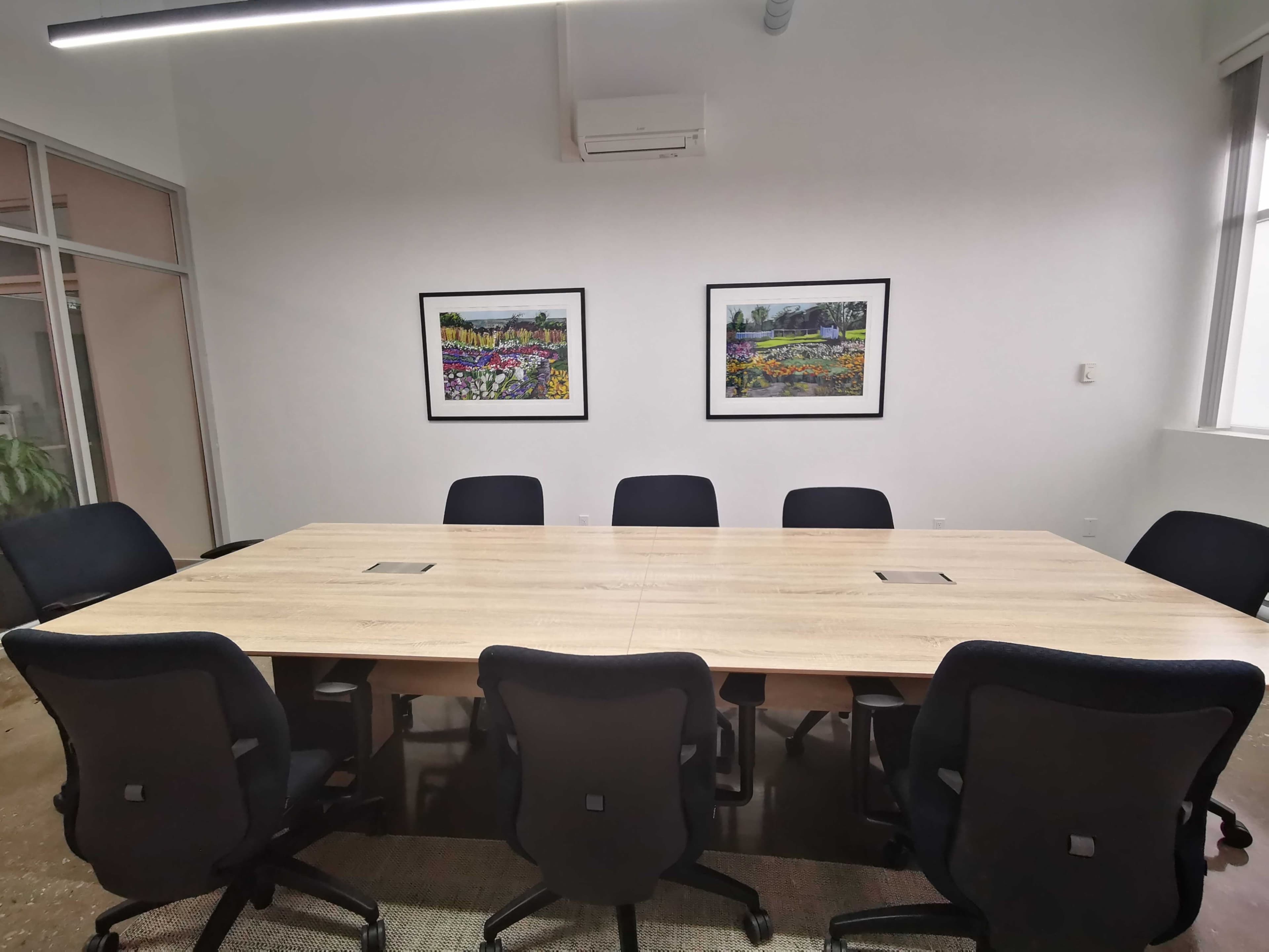 A large wooden conference table is surrounded by black office chairs in a bright meeting room, featuring two framed artwork pieces on the wall.