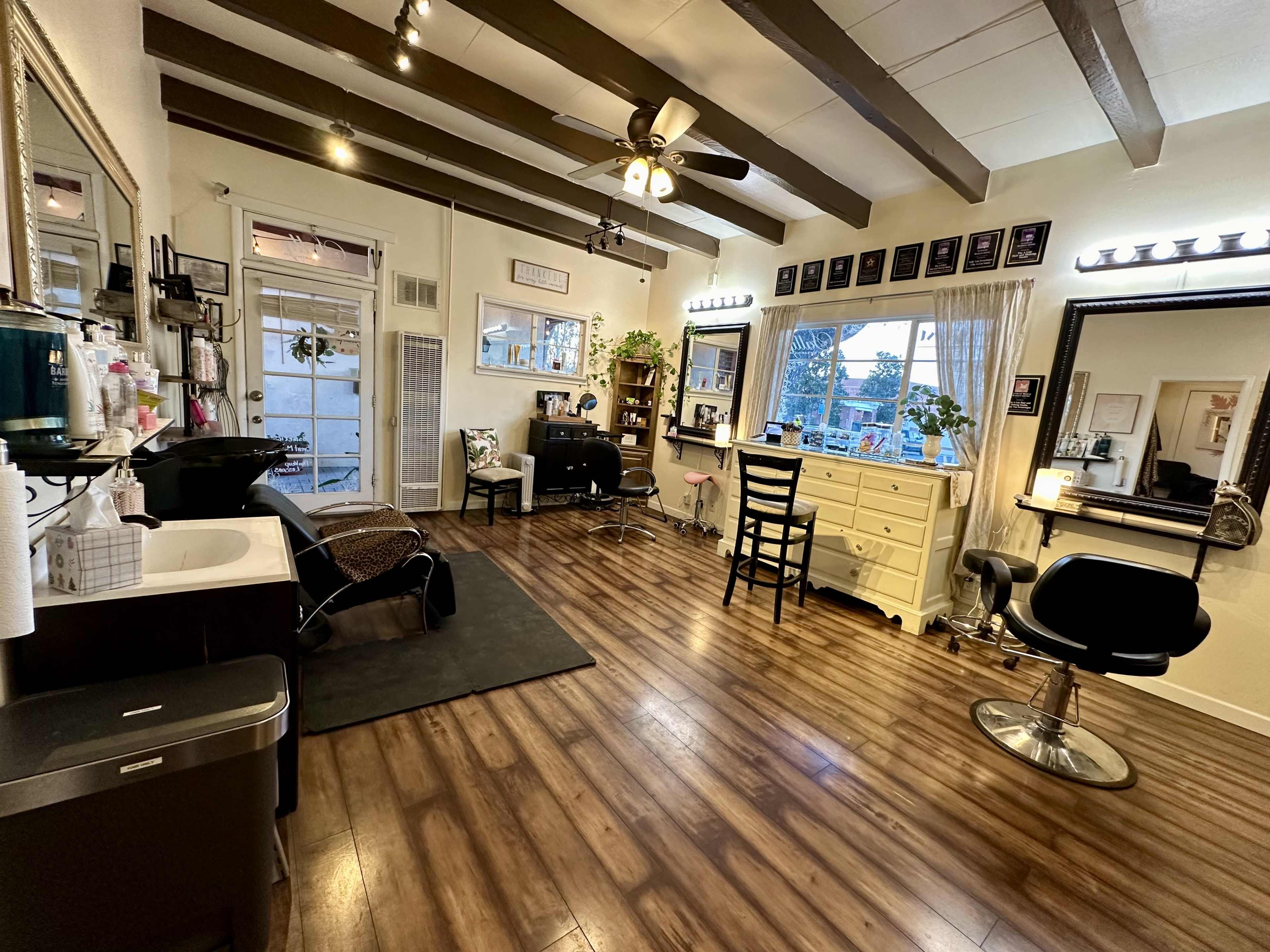 The image shows a well-lit hair salon with wooden floors, two styling stations, a sink, and various decor items on the walls.