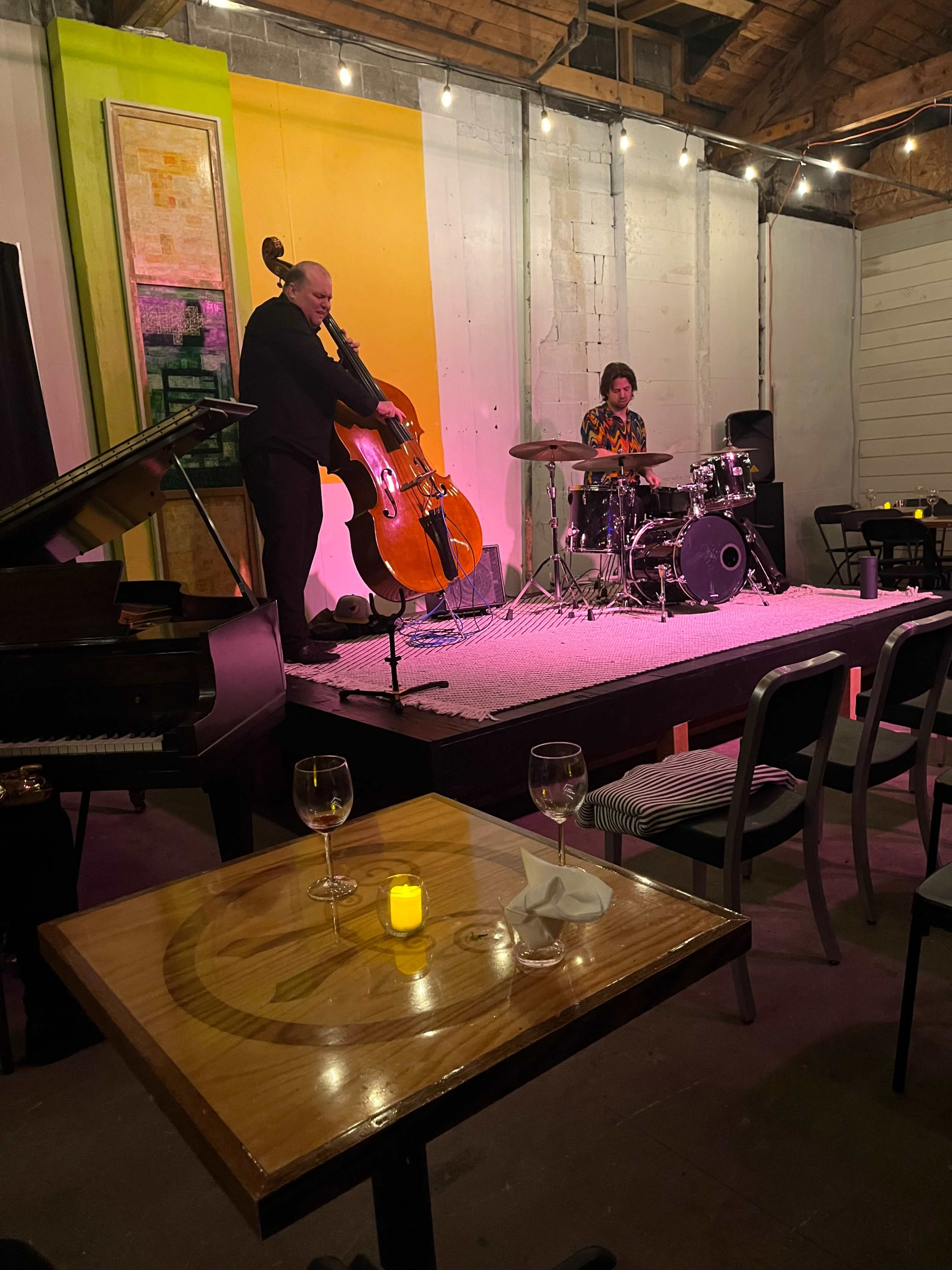 A bassist and a drummer perform on a small stage in a dimly lit venue with empty tables and glasses in the foreground.