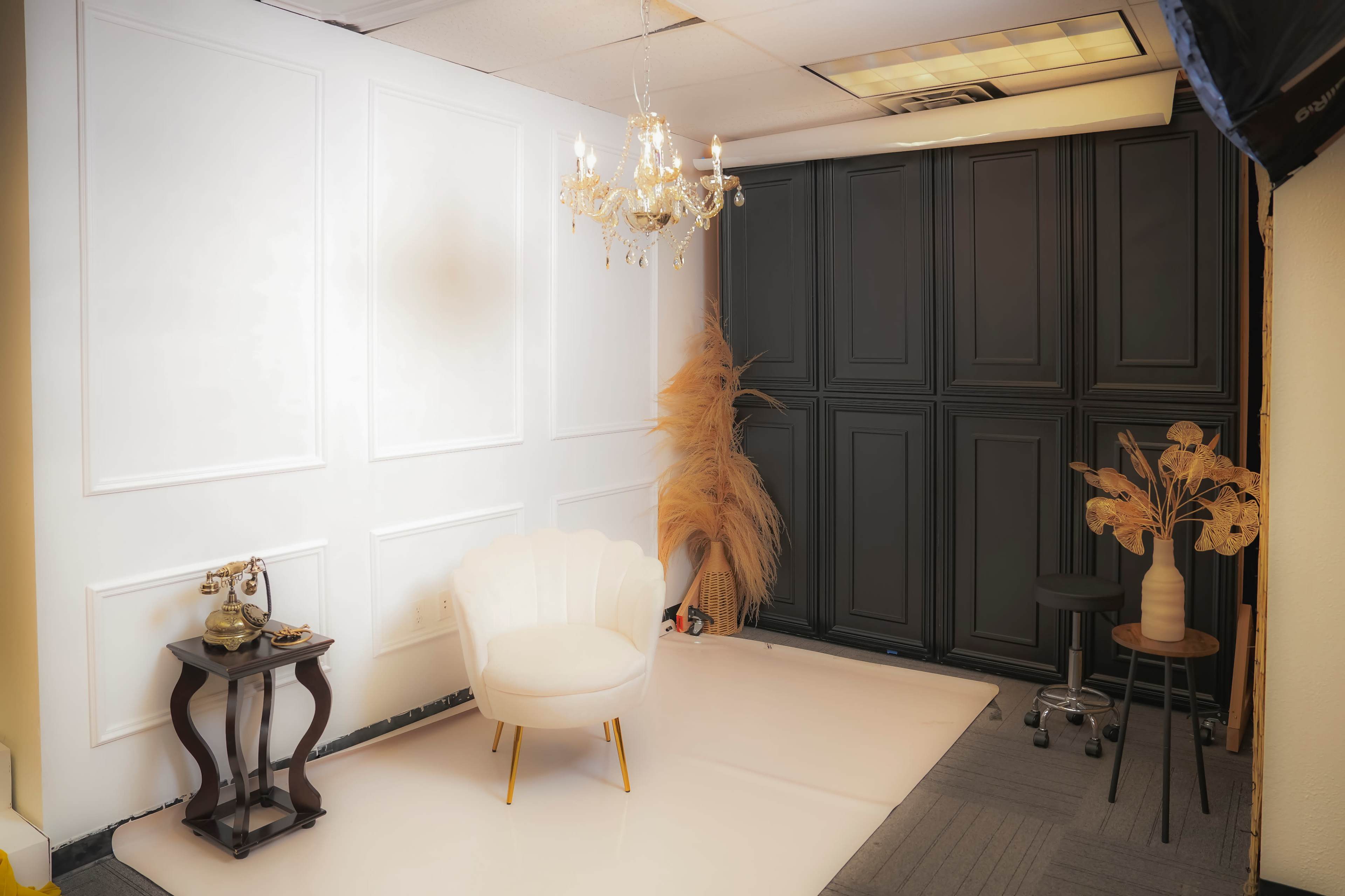 The image shows a well-decorated indoor space featuring a white wall with molding, a small chandelier, a plush white chair, and dark cabinets in the background.