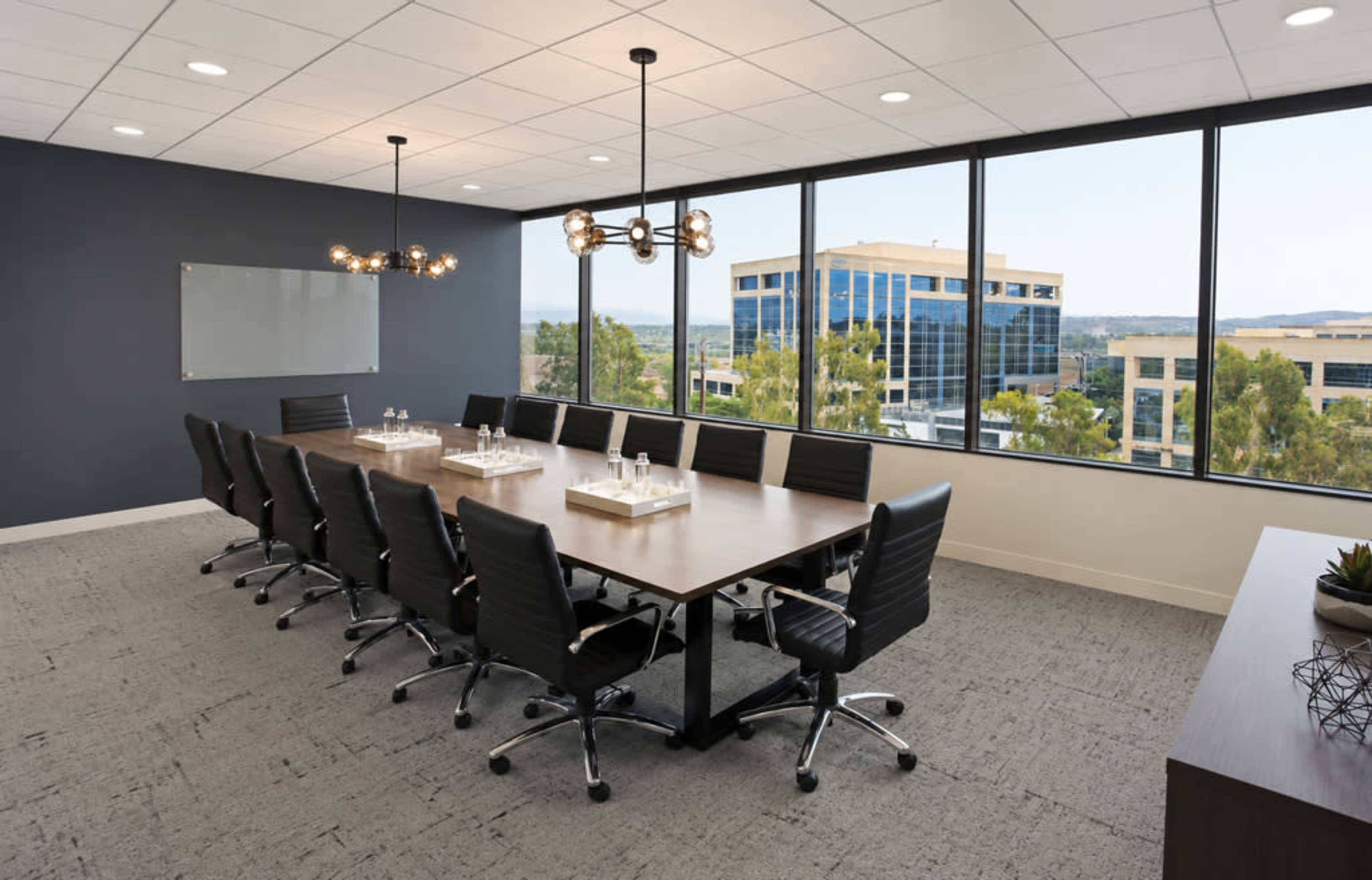 A modern conference room features a long wooden table surrounded by black ergonomic chairs and large windows providing a view of office buildings and greenery outside.