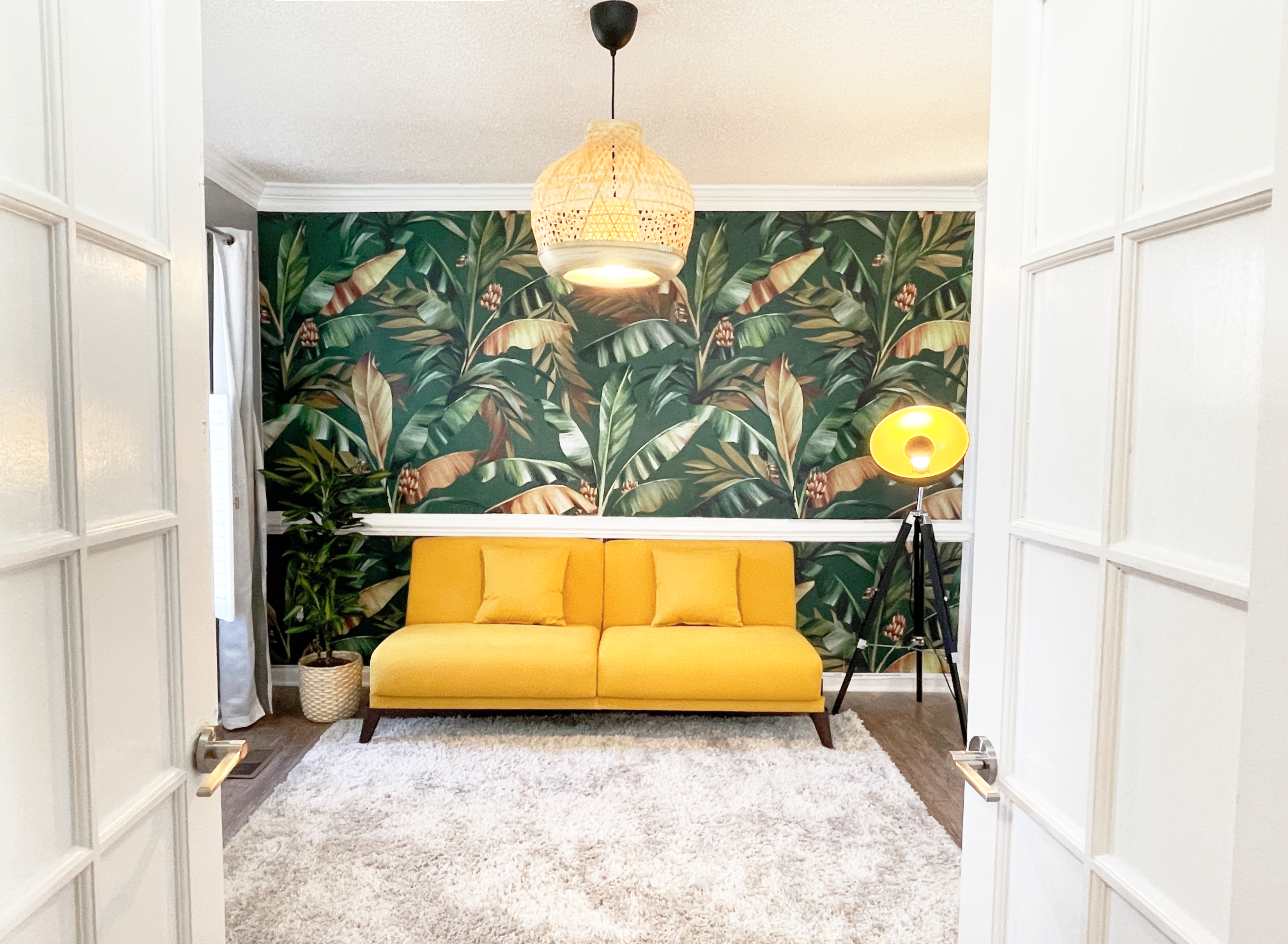 The image shows a room with a yellow sofa, a patterned green wallpaper featuring tropical leaves, and a modern lamp, viewed through glass double doors.