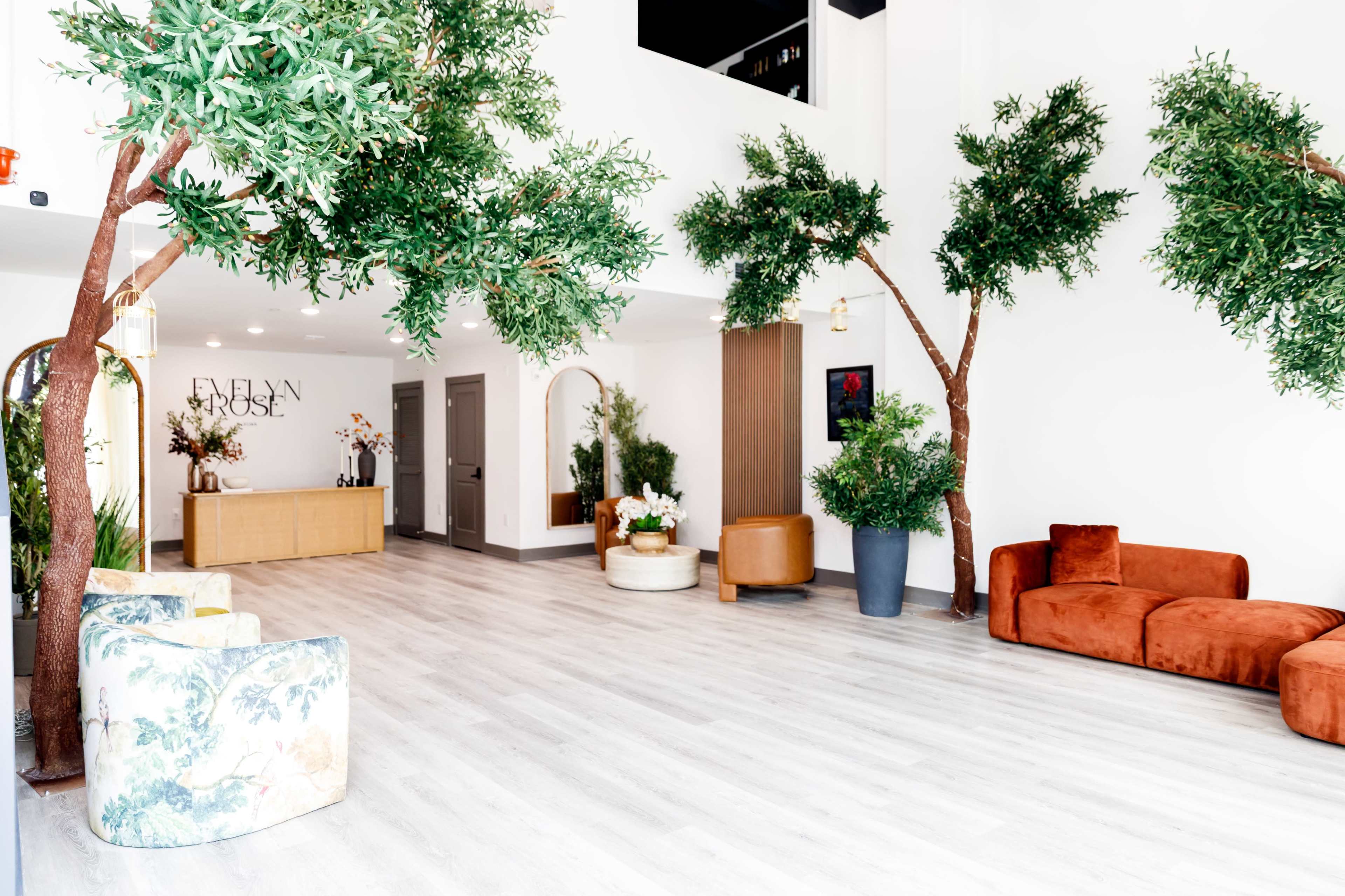 The image shows a modern indoor lobby with faux trees, a wooden reception desk, and various seating arrangements, including a curved orange sofa and patterned armchairs.