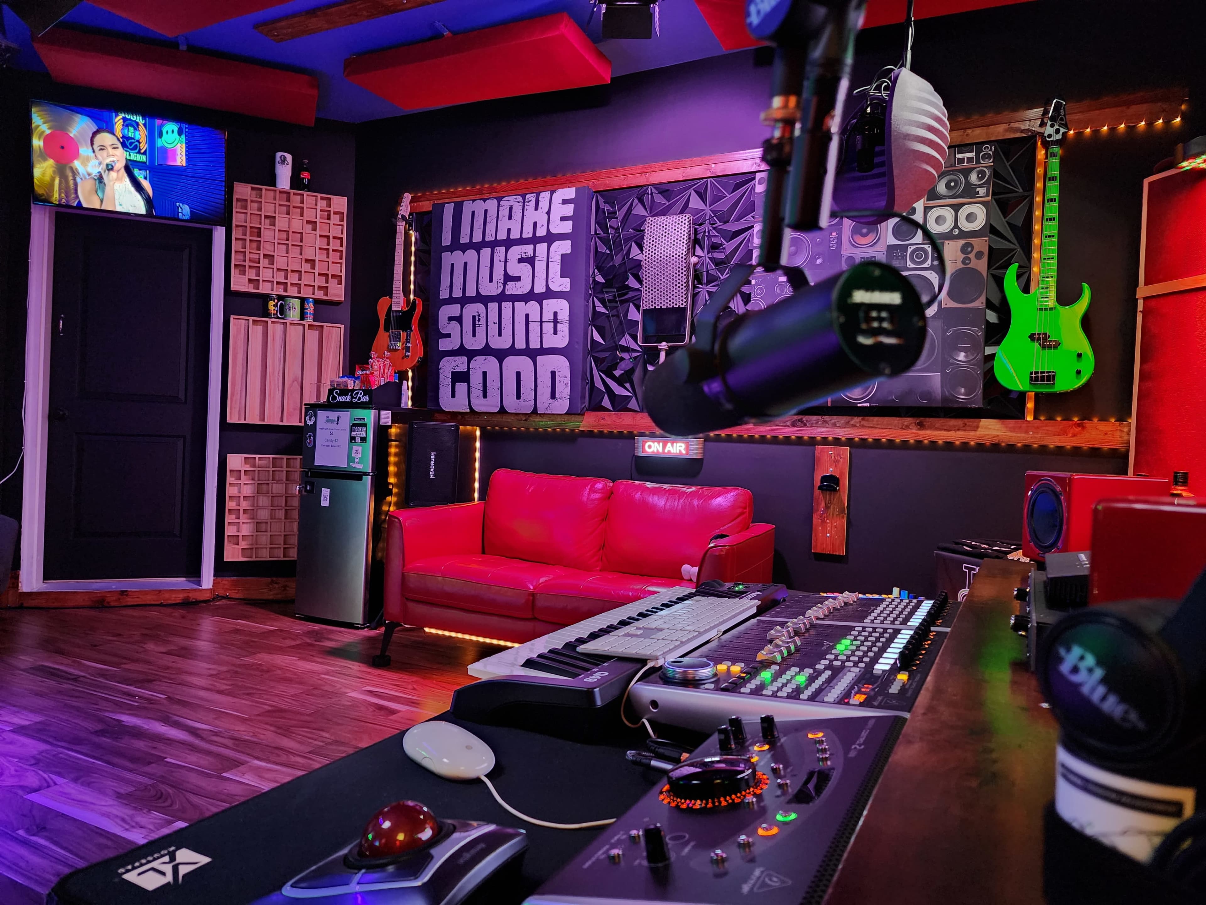 A music studio featuring a mixing console, a red couch, green guitars on the wall, and a television displaying a music-themed program.