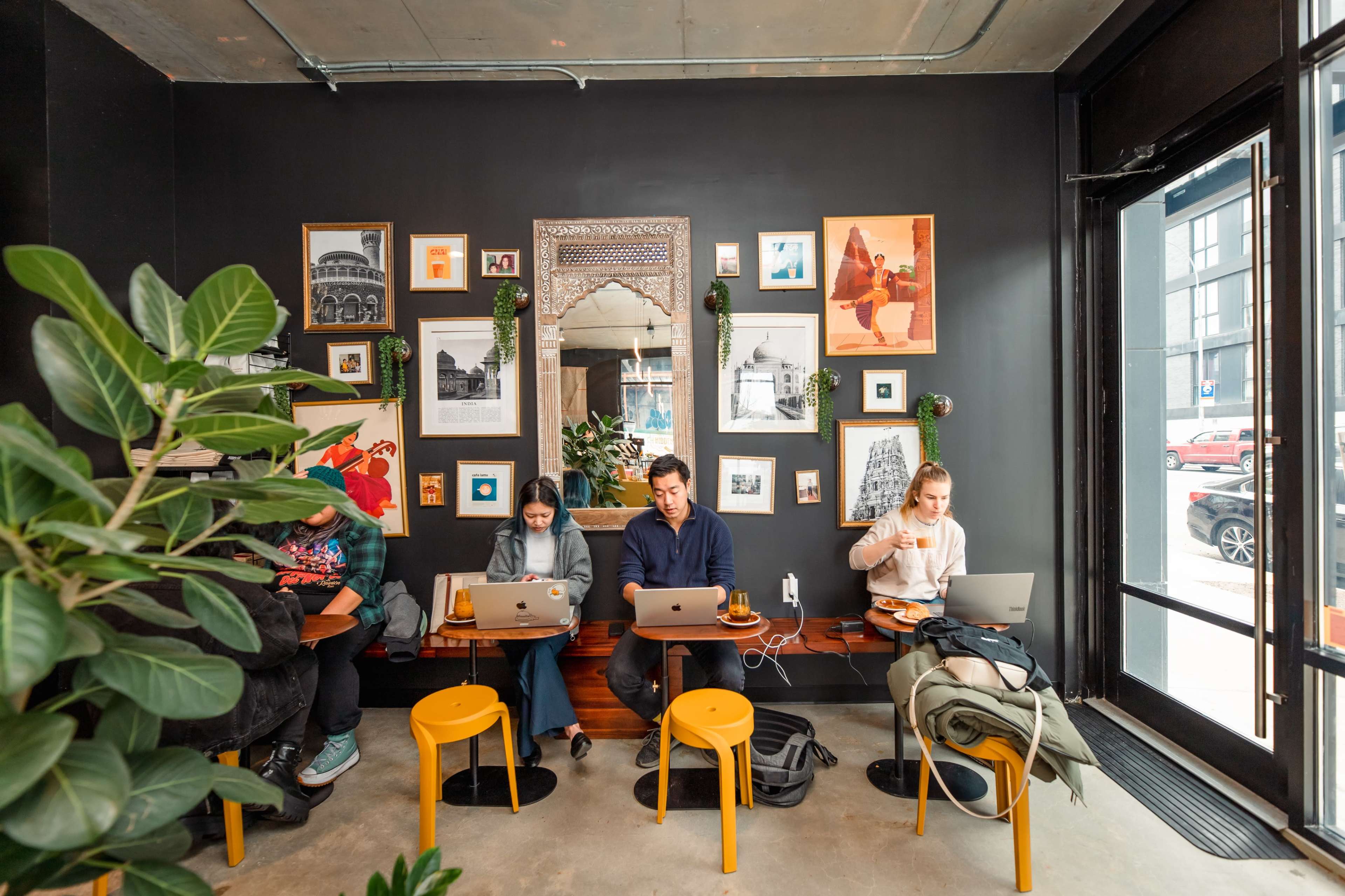 Three people work at tables in a café, surrounded by framed artwork and plants, with a glass door allowing visibility to the exterior.
