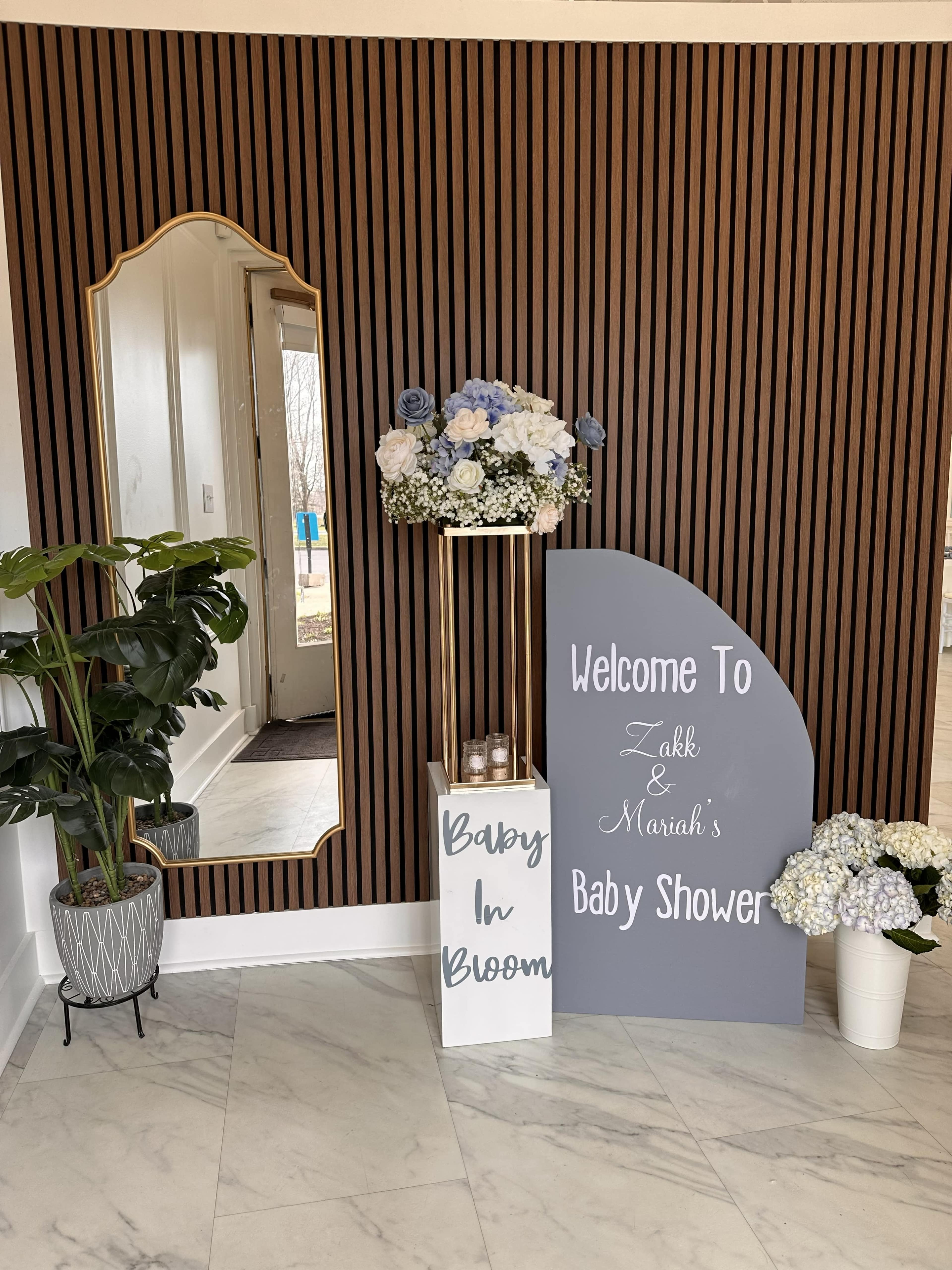 The image shows a decorated entryway for a baby shower, featuring a mirror, a floral arrangement, and signs welcoming guests, set against a striped wall.