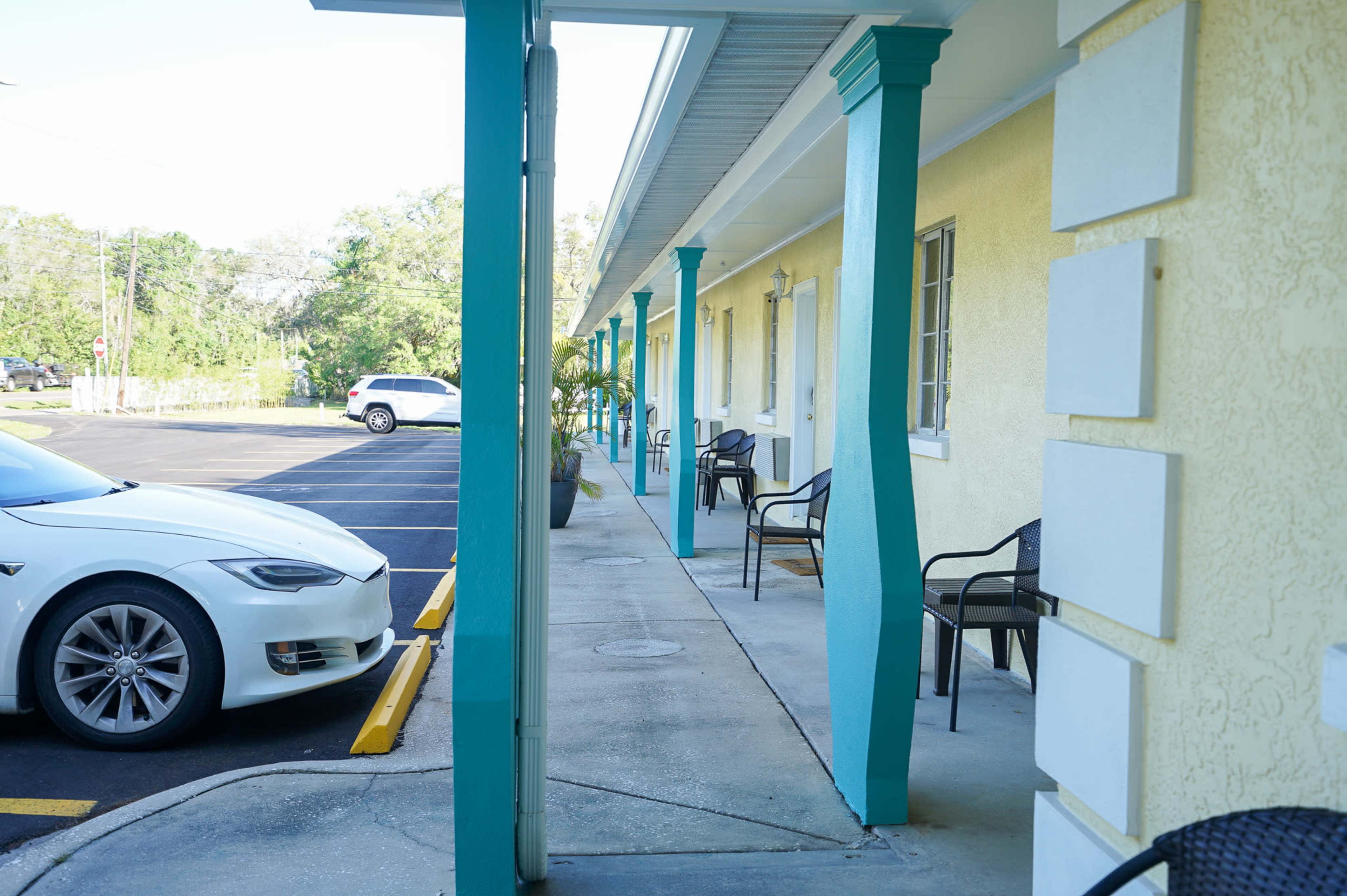A white car is parked in front of a motel with turquoise columns and seating along the walkway.