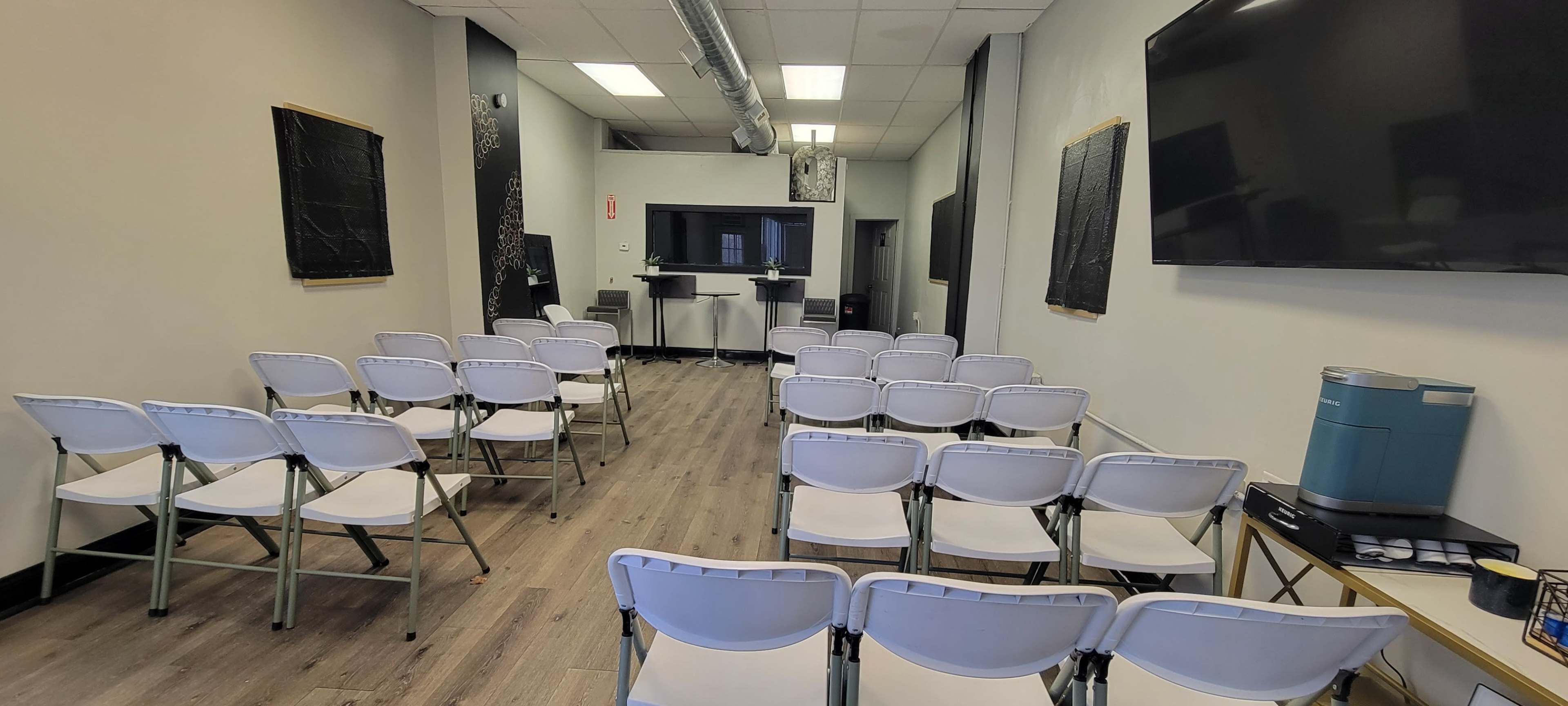 The image shows a training or meeting room arranged with white folding chairs facing a presentation area, featuring a large screen and a small table.