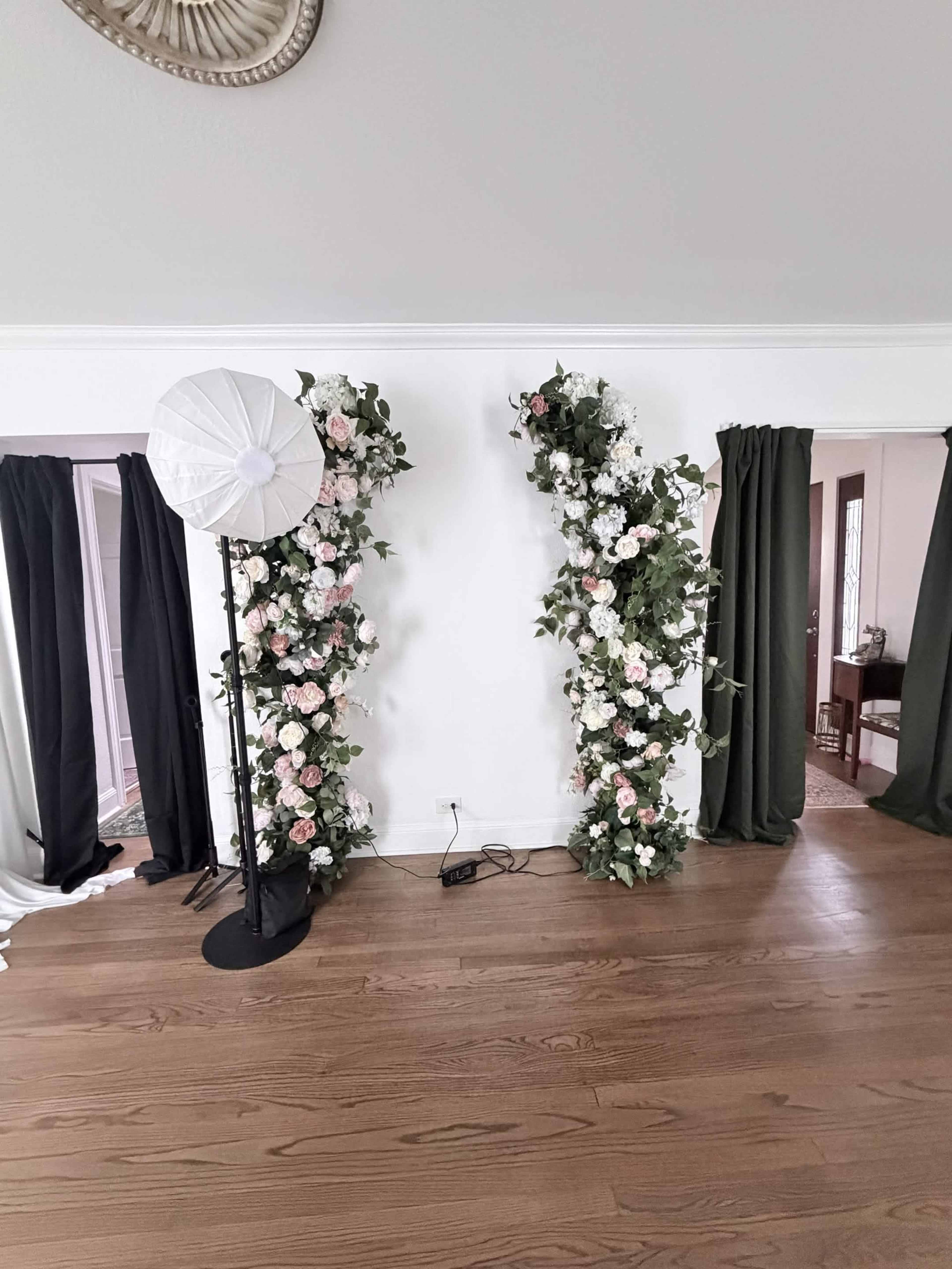 Two floral arrangements with pink and white flowers are positioned against a white wall, flanking a black photography light stand on a wooden floor.